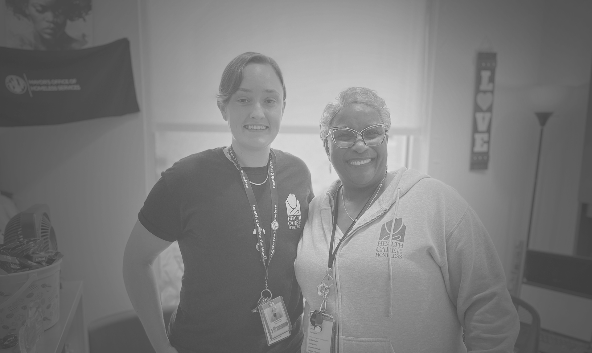 A light-skinned person with short medium-toned hair stands in an office next to a medium-skinned woman with glasses and short light hair.