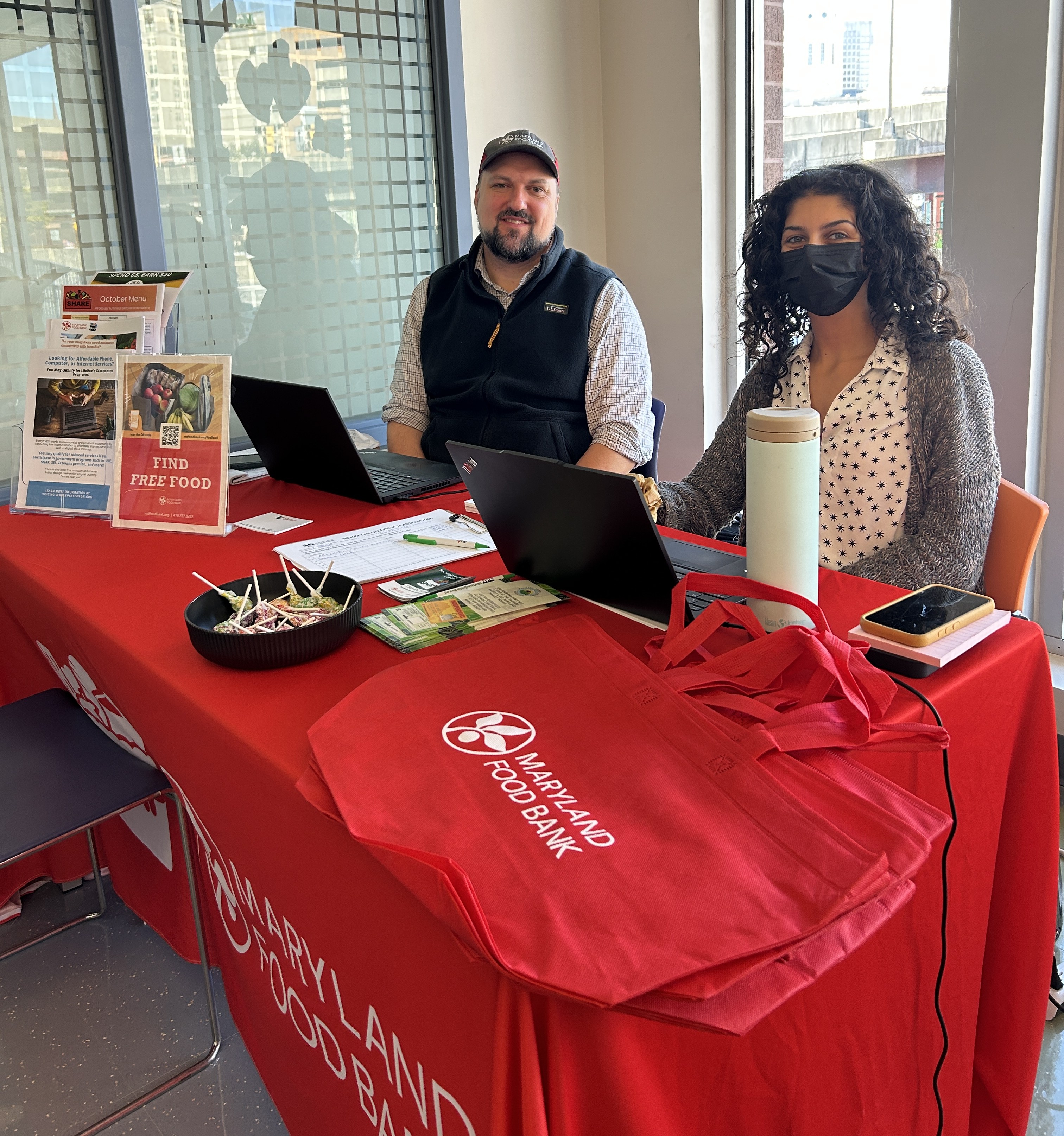 A light-skinned man in a hat sits next to a light-skinned woman wearing a black mask at a table with a red tablecloth.