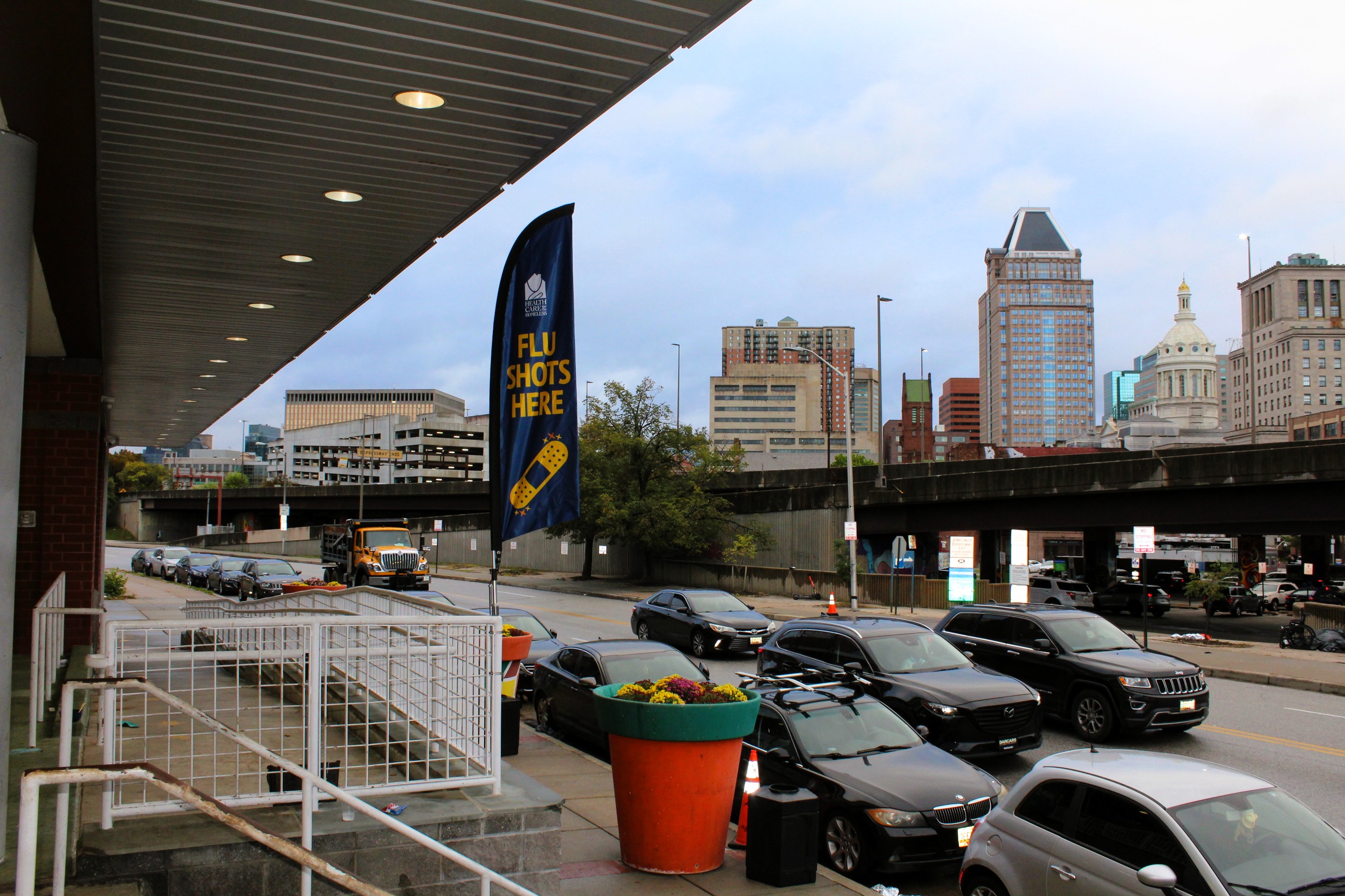 A view of the Baltimore City skyline. A blue and orange teardrop sign reads, "Flu shots here" with a bandage icon underneath.