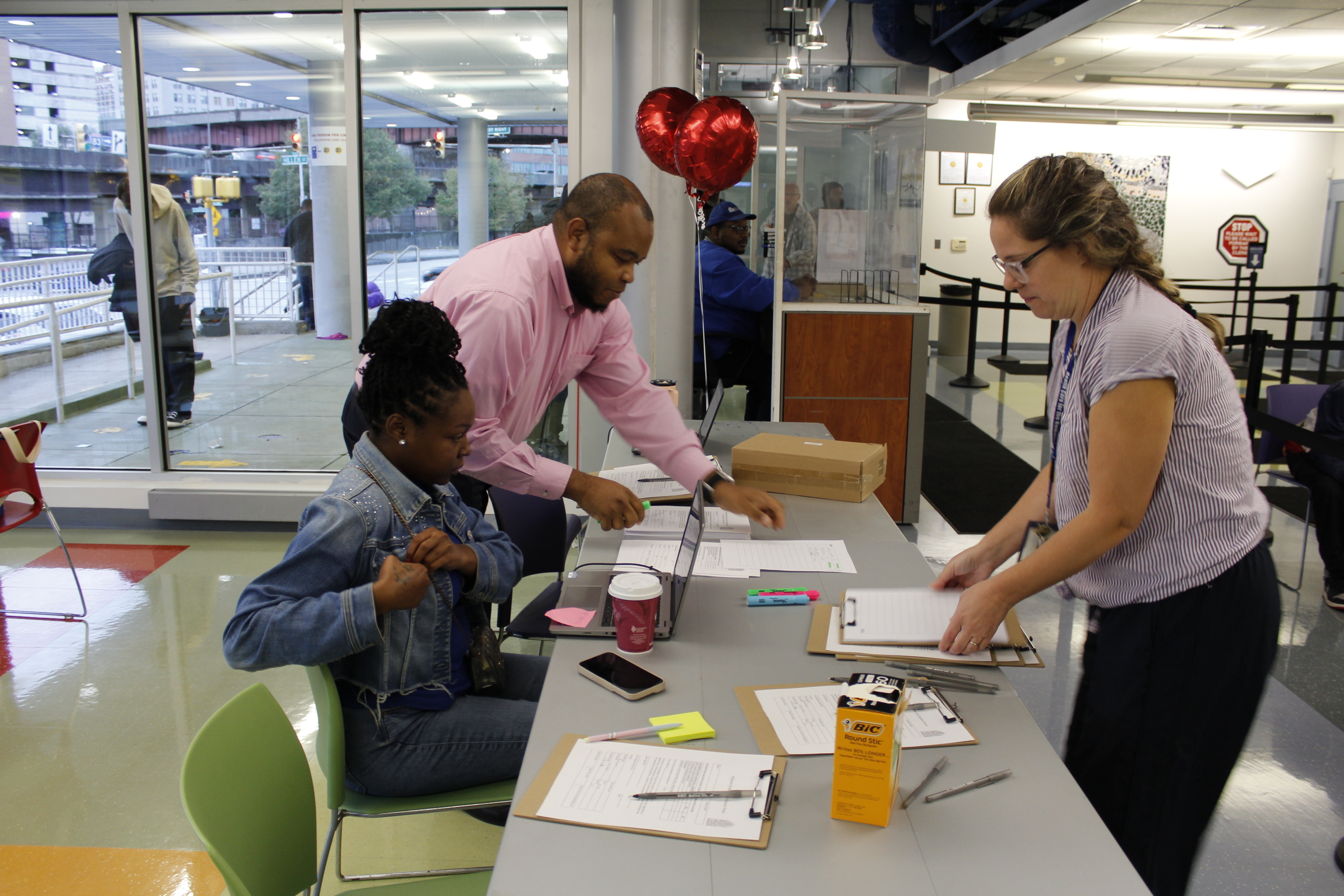 A light-skinned woman with blond hair stands in front of a registration table, facing a sitting dark-skinned woman with dark hair and a standing medium-skinned man in a pink shirt.