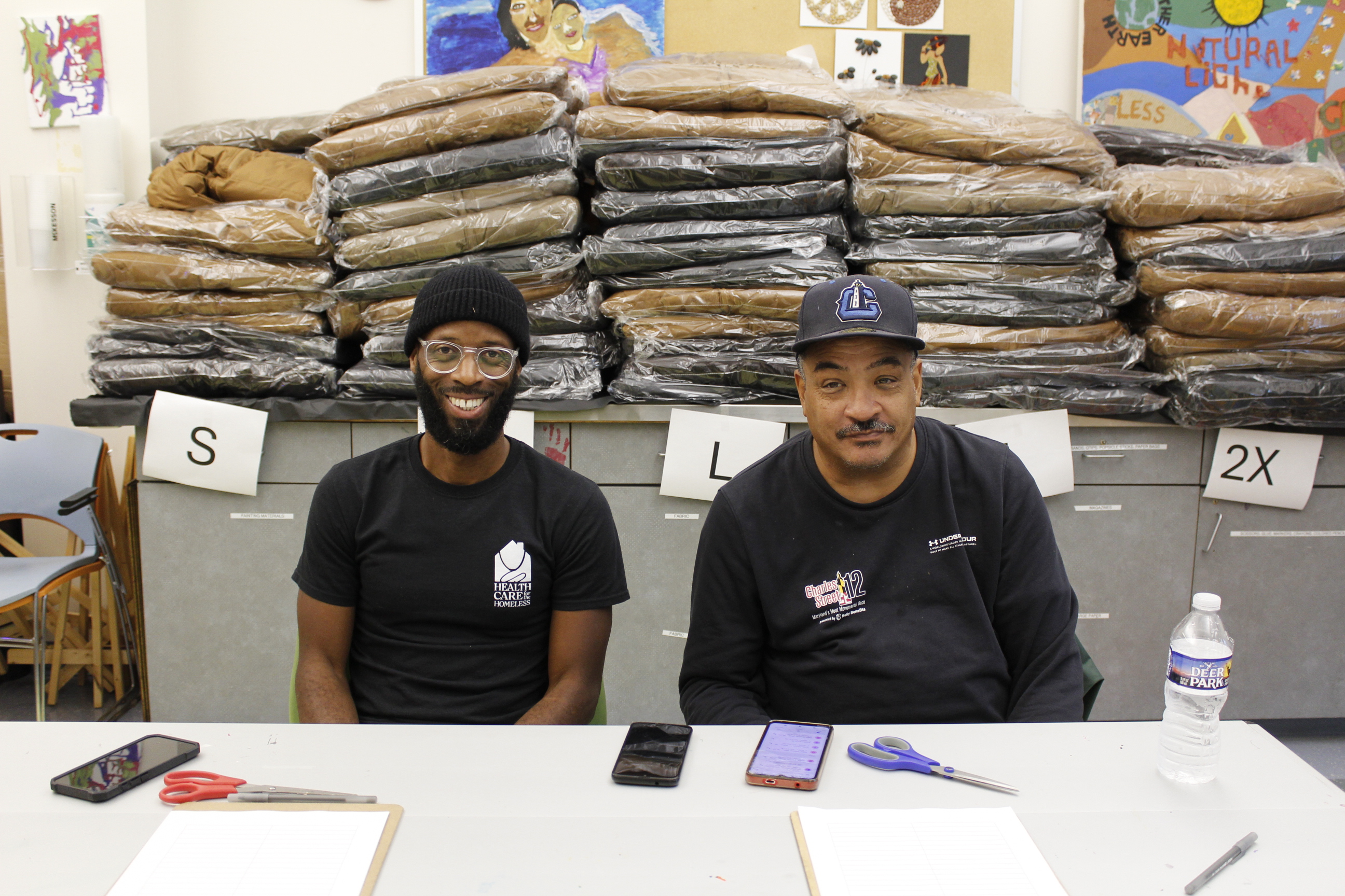 A dark-skinned man with a dark beanie sits at a table next to a dark-skinned man with a baseball hat. Behind them are piles of folded coats on a counter.