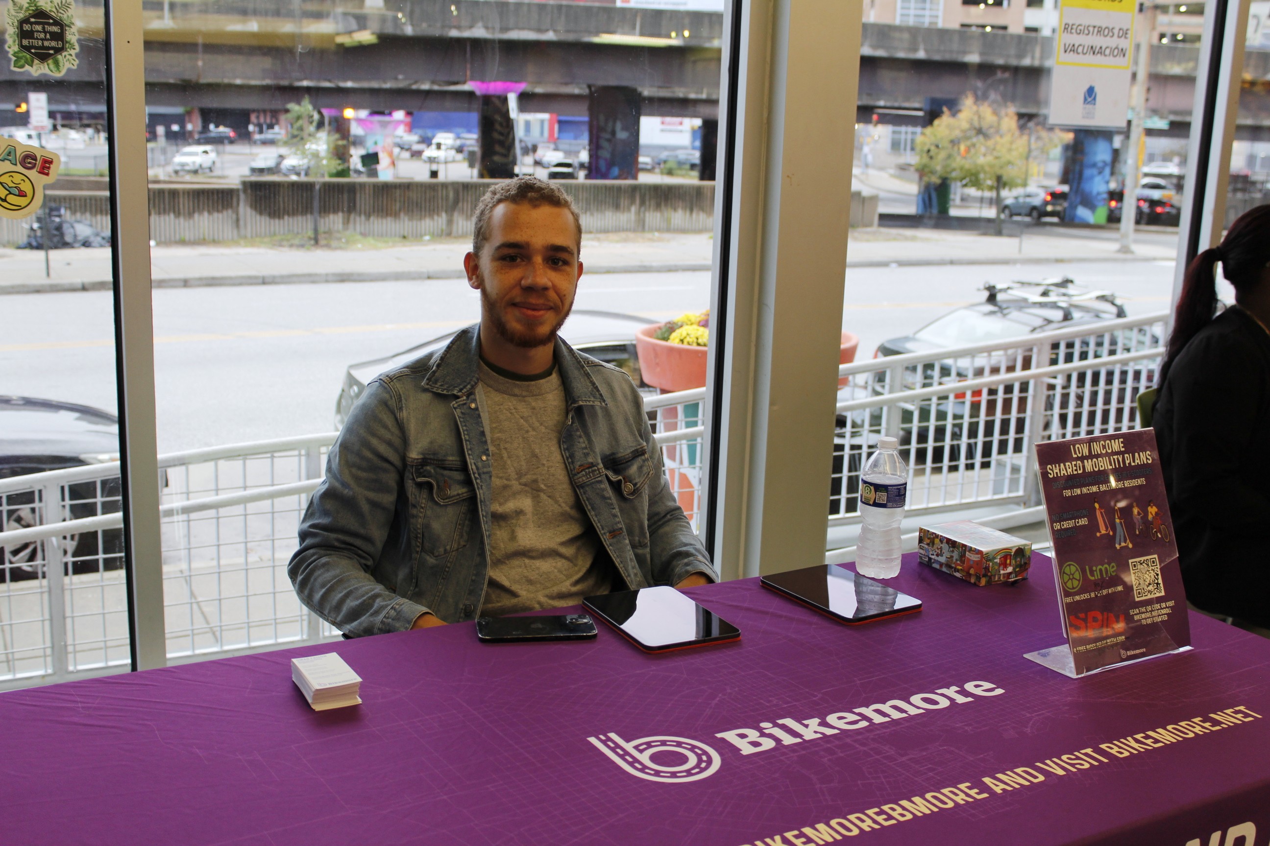 A medium-skinned man sitting at a table with a purple tablecloth that reads, "Bikemore."