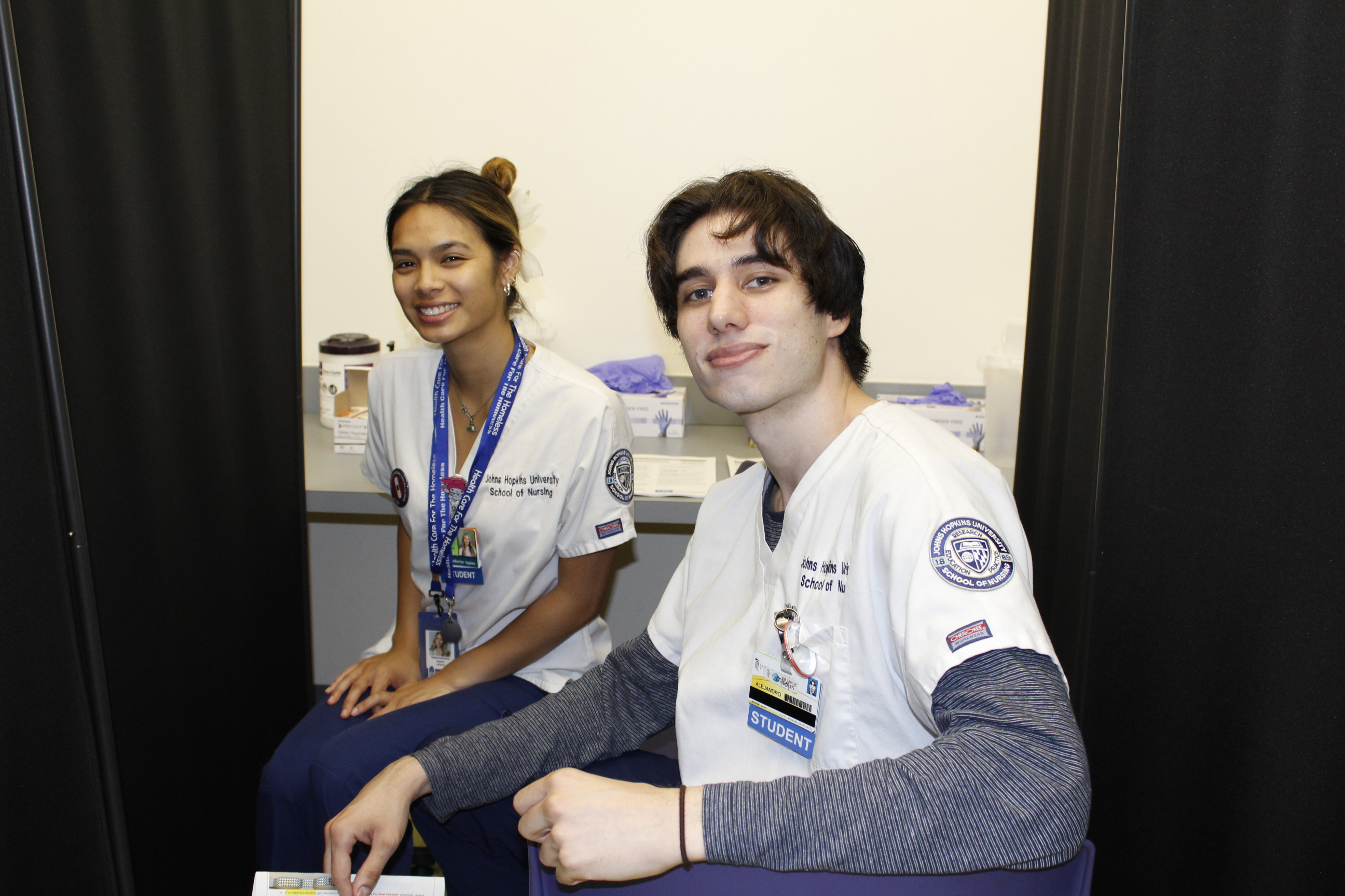 A medium-skinned nursing student and a light-skinned nursing student are seating and smiling. They both wear white scrub tops.