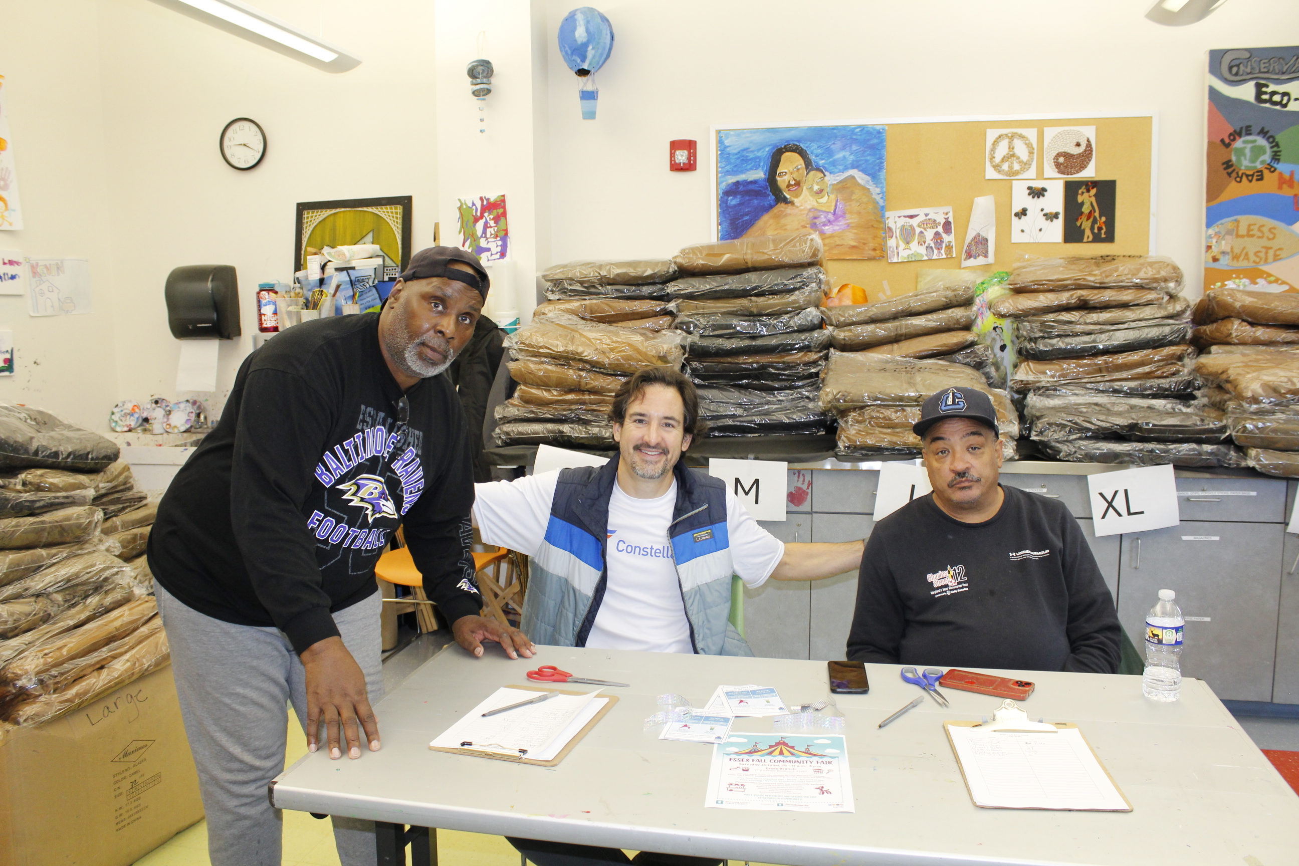 A dark-skinned man stands next to a table with a light-skinned man and a medium-skinned man. Behind them are piles of coats on a cabinet countertop.
