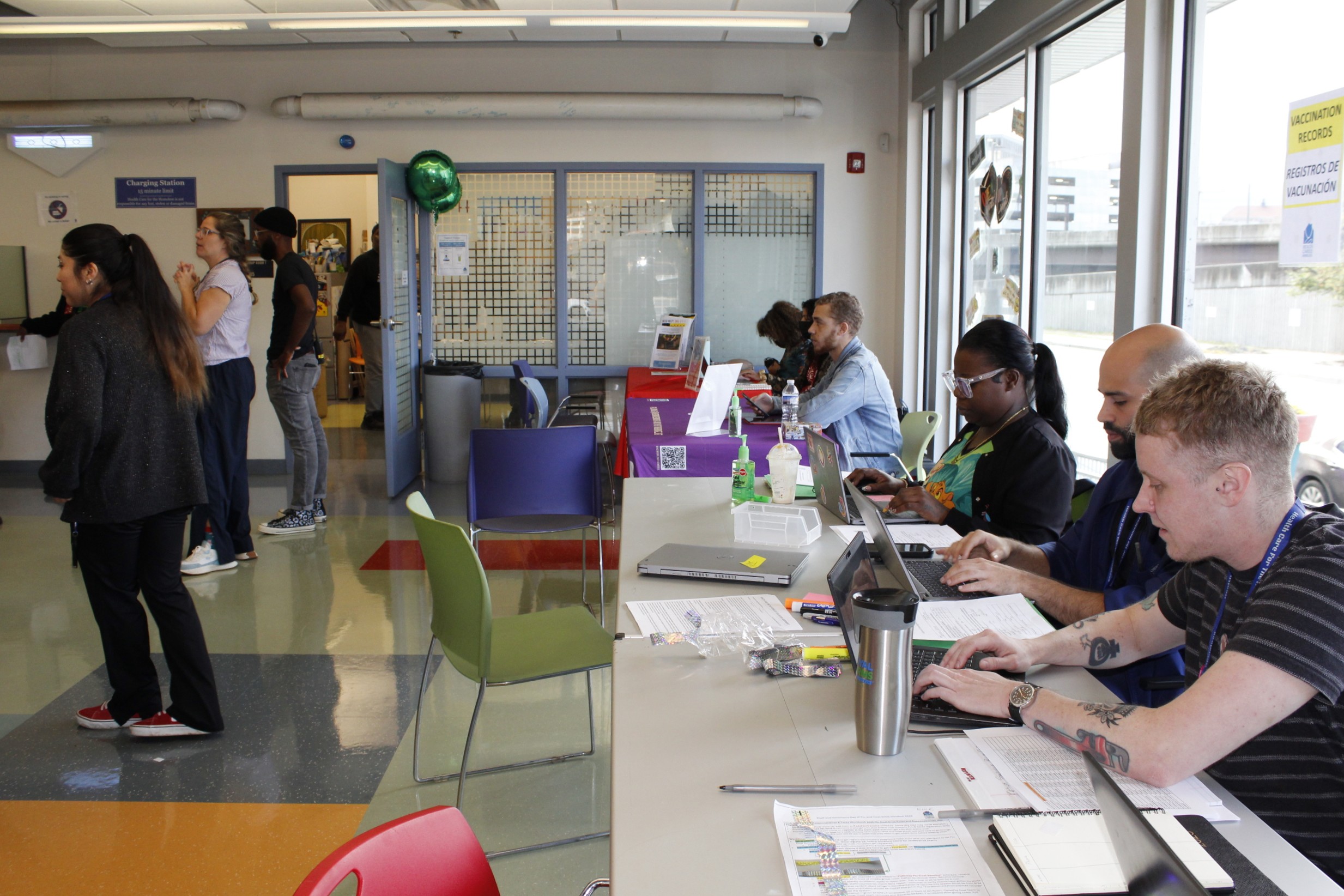 Some staff sit at the vaccination records table while others observe the clients waiting in the lobby.