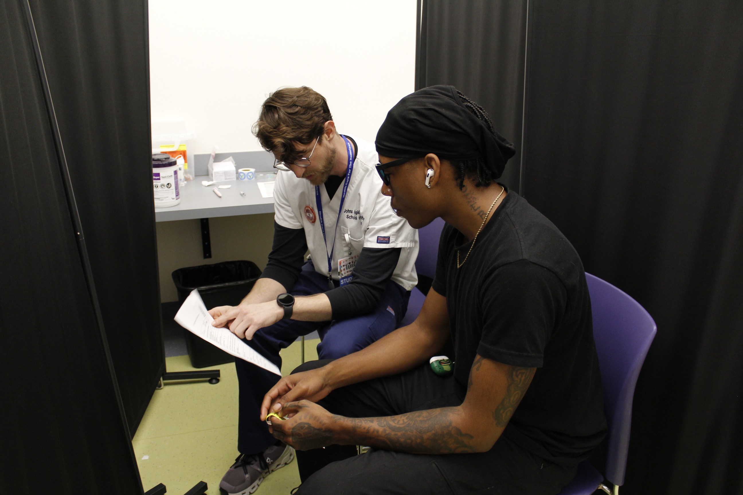 A light-skinned nurse wearing a white scrub top advises a dark-skinned client. Both are seated in a curtained booth.