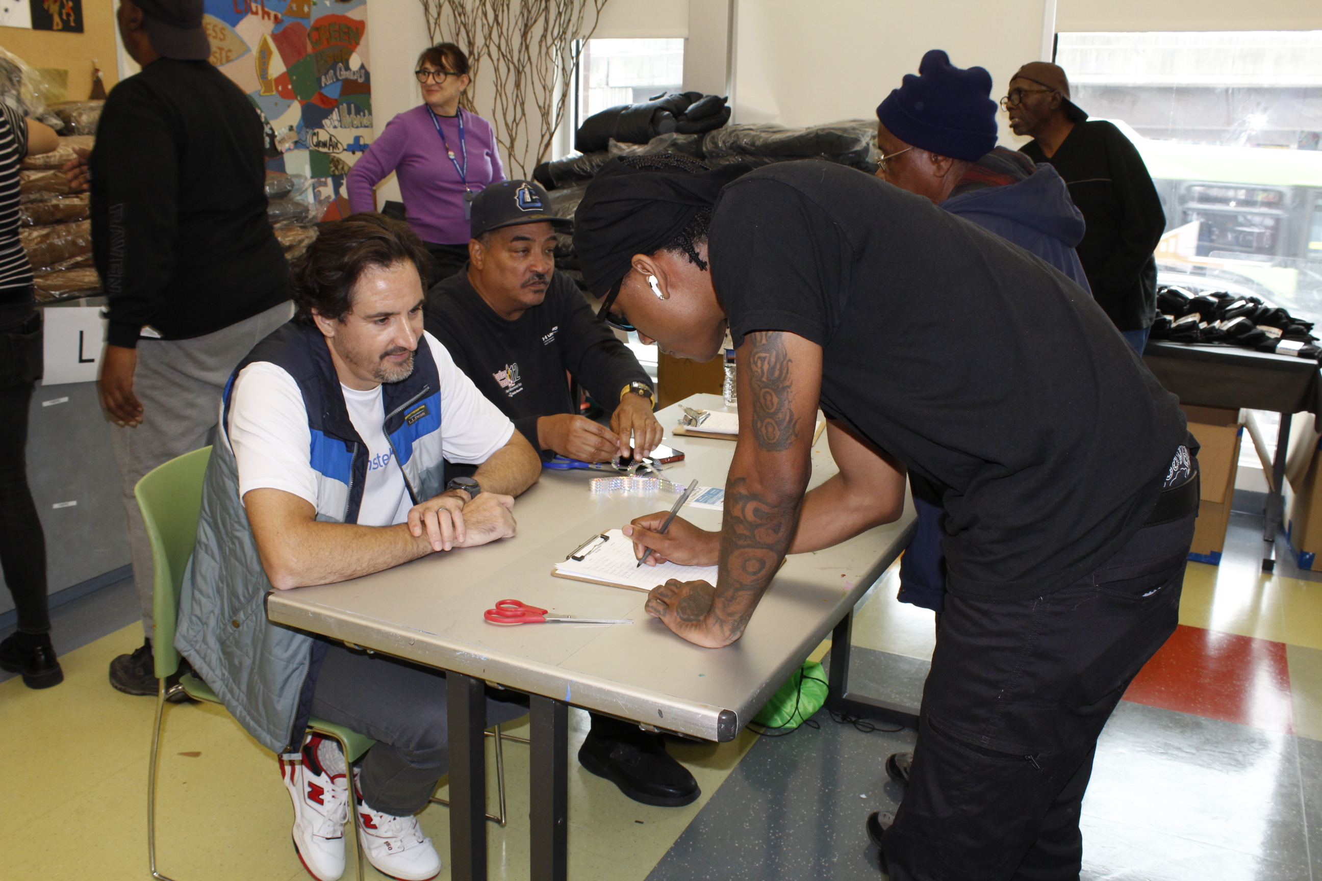 A dark-skinned man wearing dark clothing signs for his coat. A light-skinned man wearing a blue vest sits at the table.