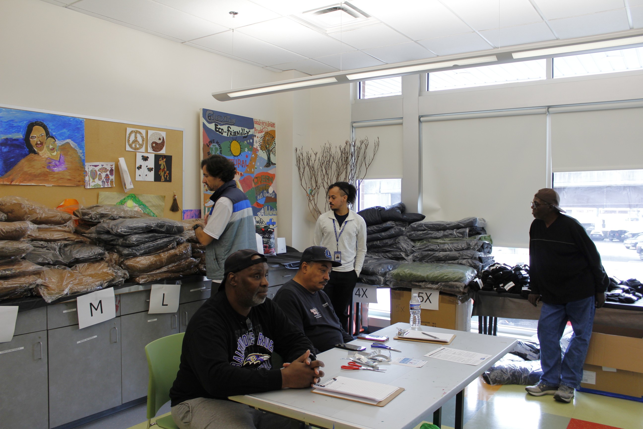 A group of people wait to hand out coats. There are coats piled on a cabinet counter and colorful artwork on the back wall.