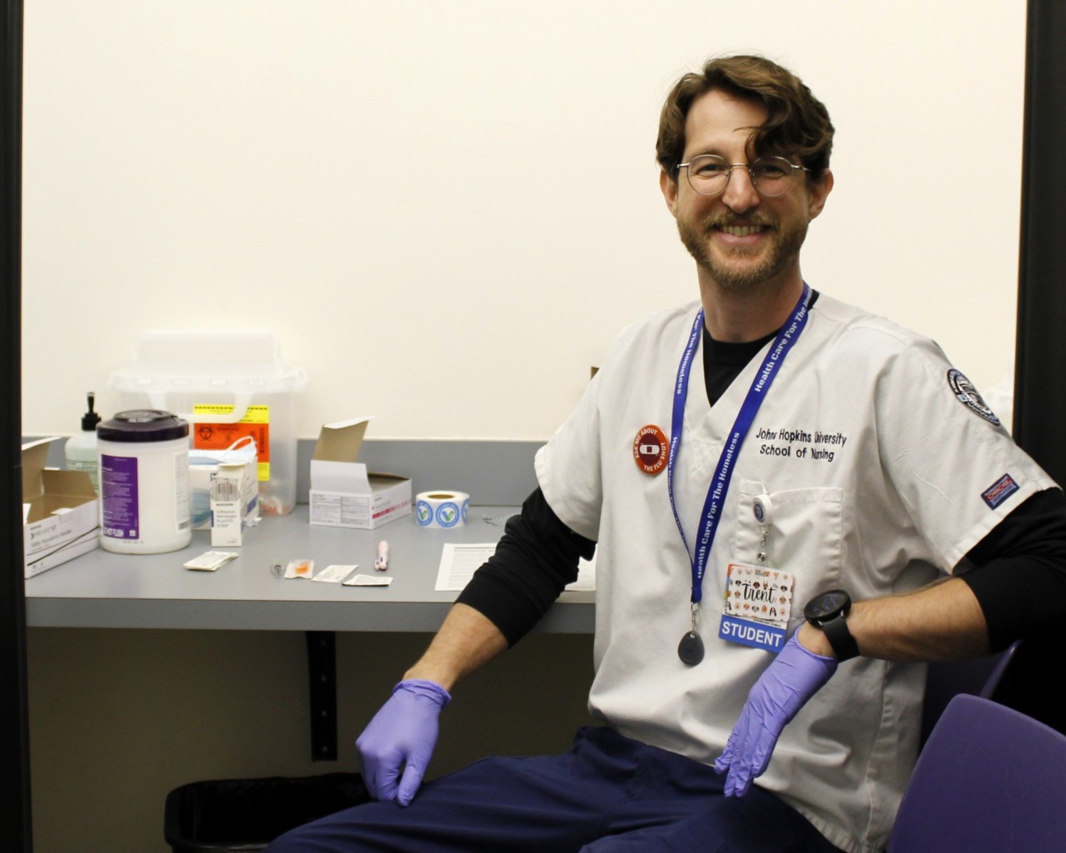 A light-skinned man wearing a white scrub top and blue gloves sits and smiles while waiting to administer flu shots.