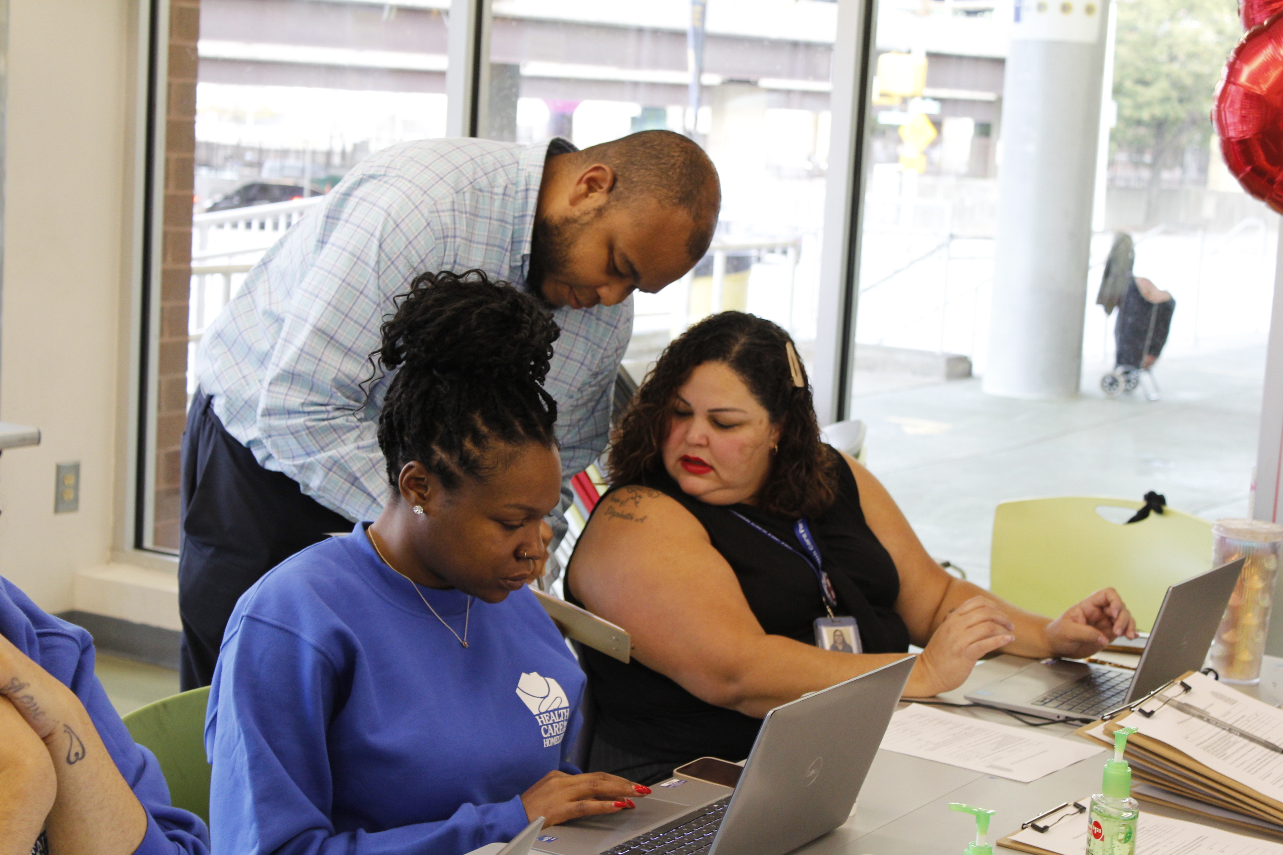 A medium-skinned man leans to speak to a sitting medium-skinned woman. A dark-skinned woman next to them types on her laptop.