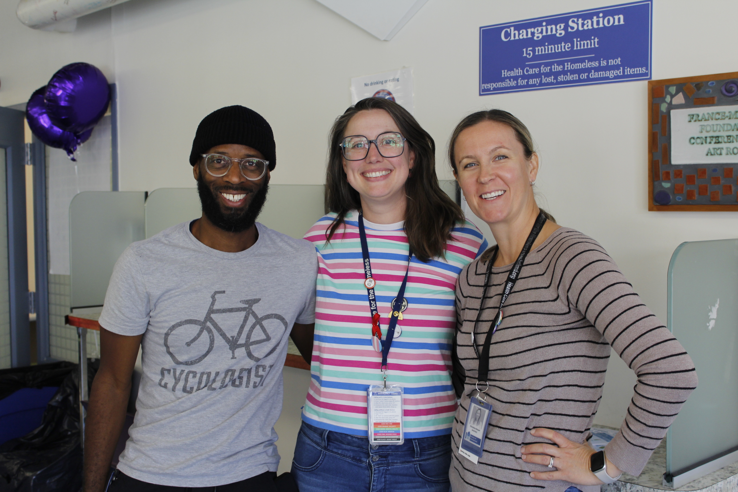 A dark-skinned man wearing a hat, a light-skinned woman with glasses, and a light-skinned woman smile for the camera.