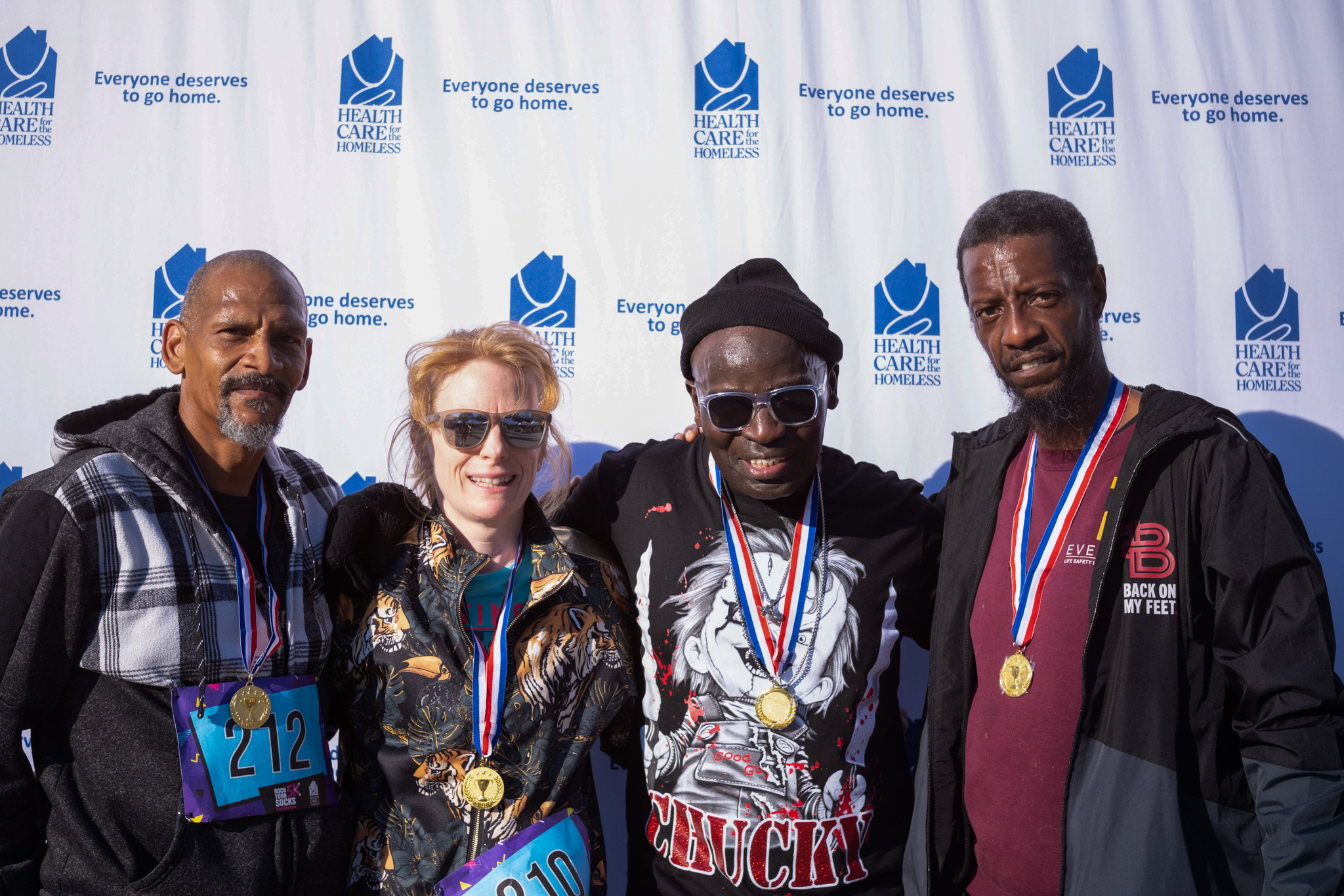 Four people in front of the Health Care for the Homeless step-and-repeat. They are wearing medals.
