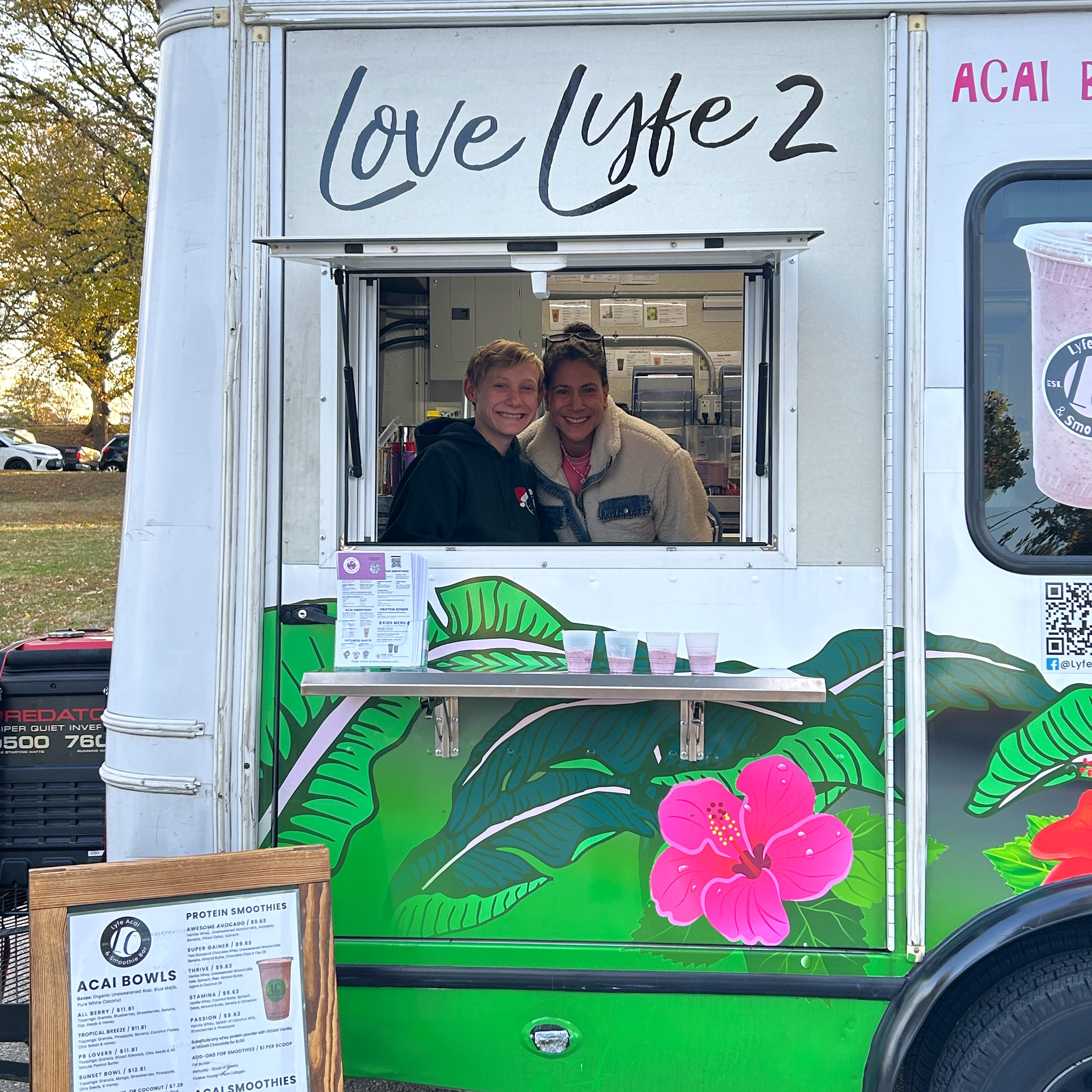 Two light-skinned people smile from the window of a food truck.