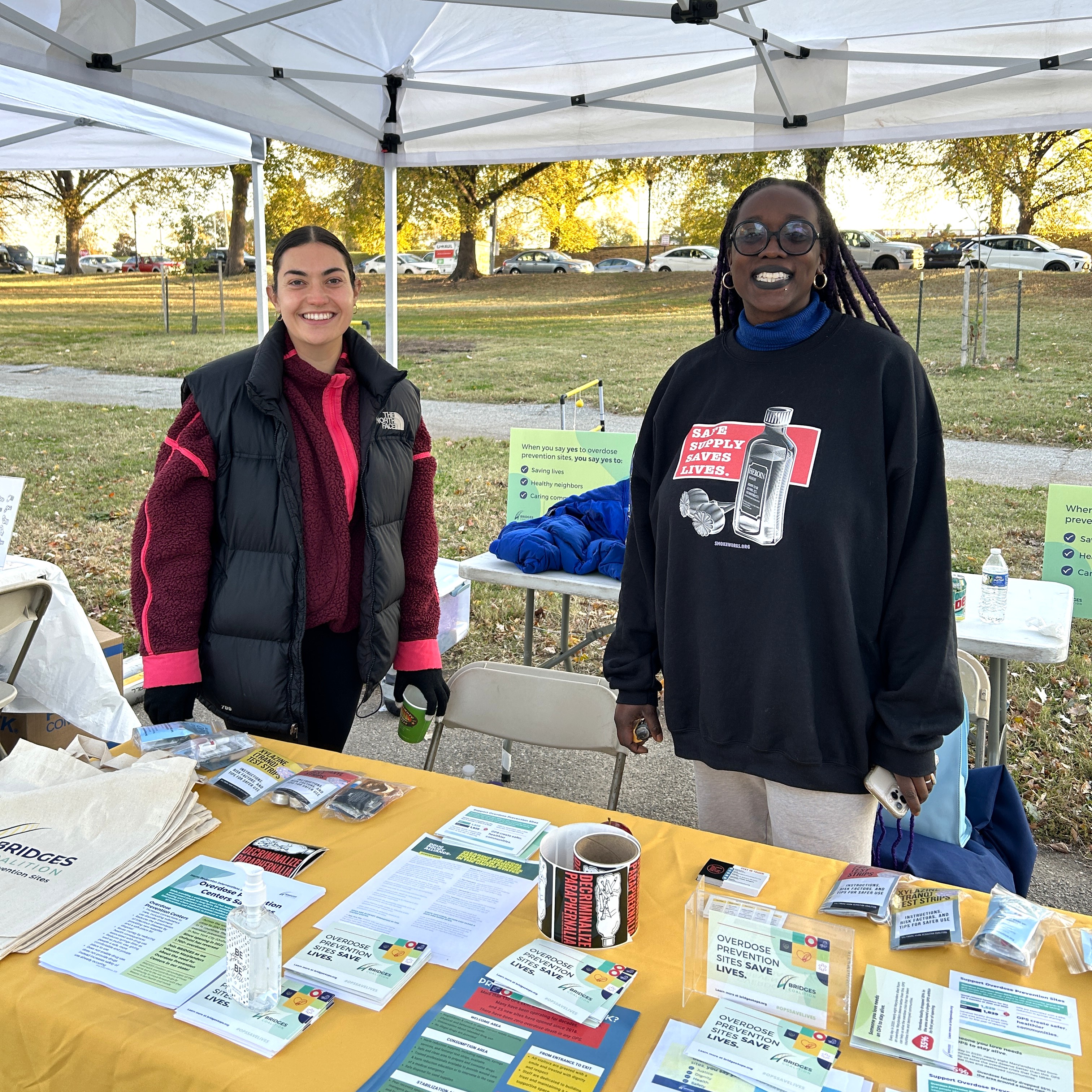 A light-skinned woman and dark-skinned woman from Baltimore Harm Reduction Coalition stand behind a table with a yellow tablecloth.