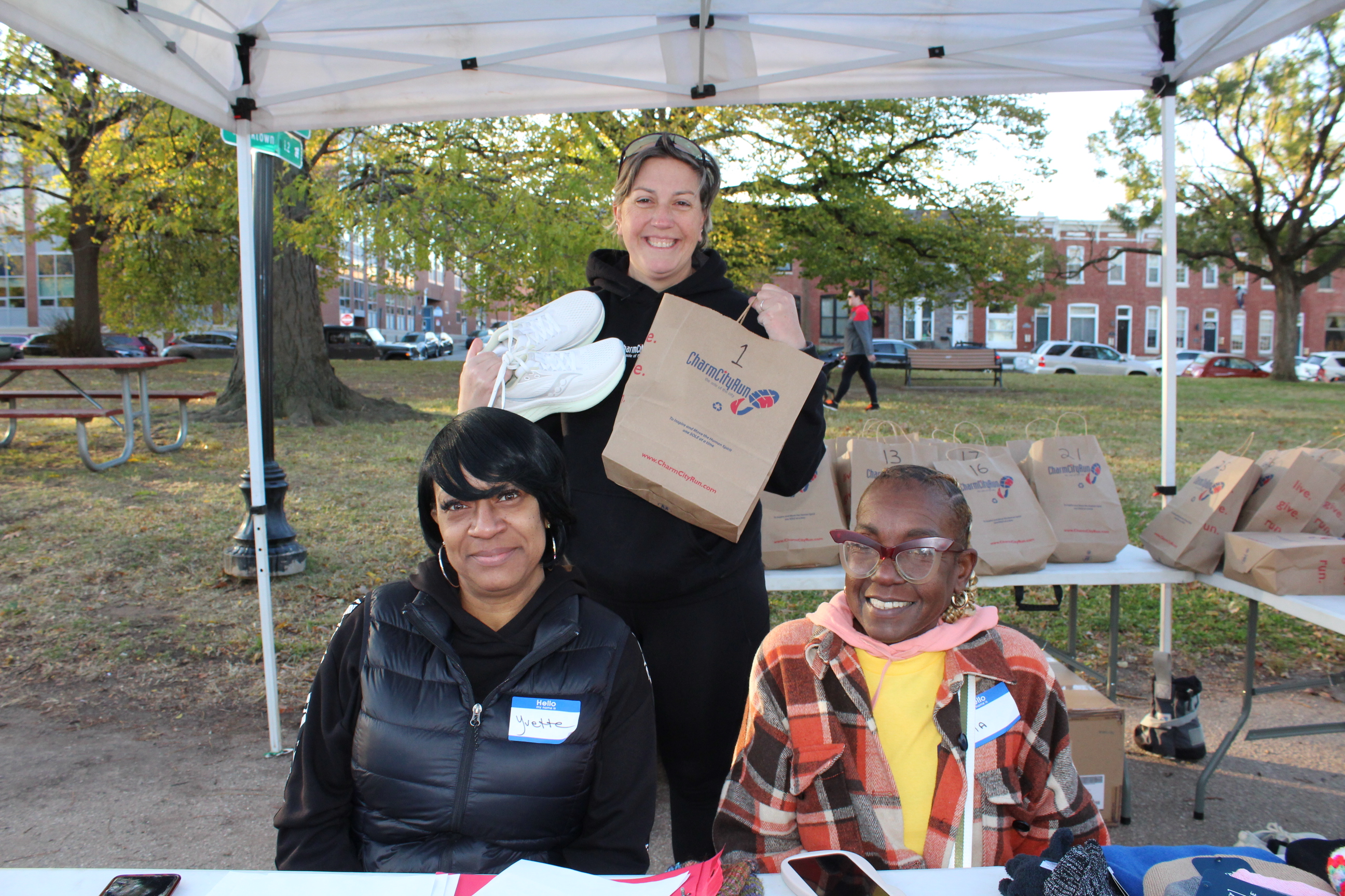 Two people sit under a tent at a table while another person hold a brown bag up behind them.
