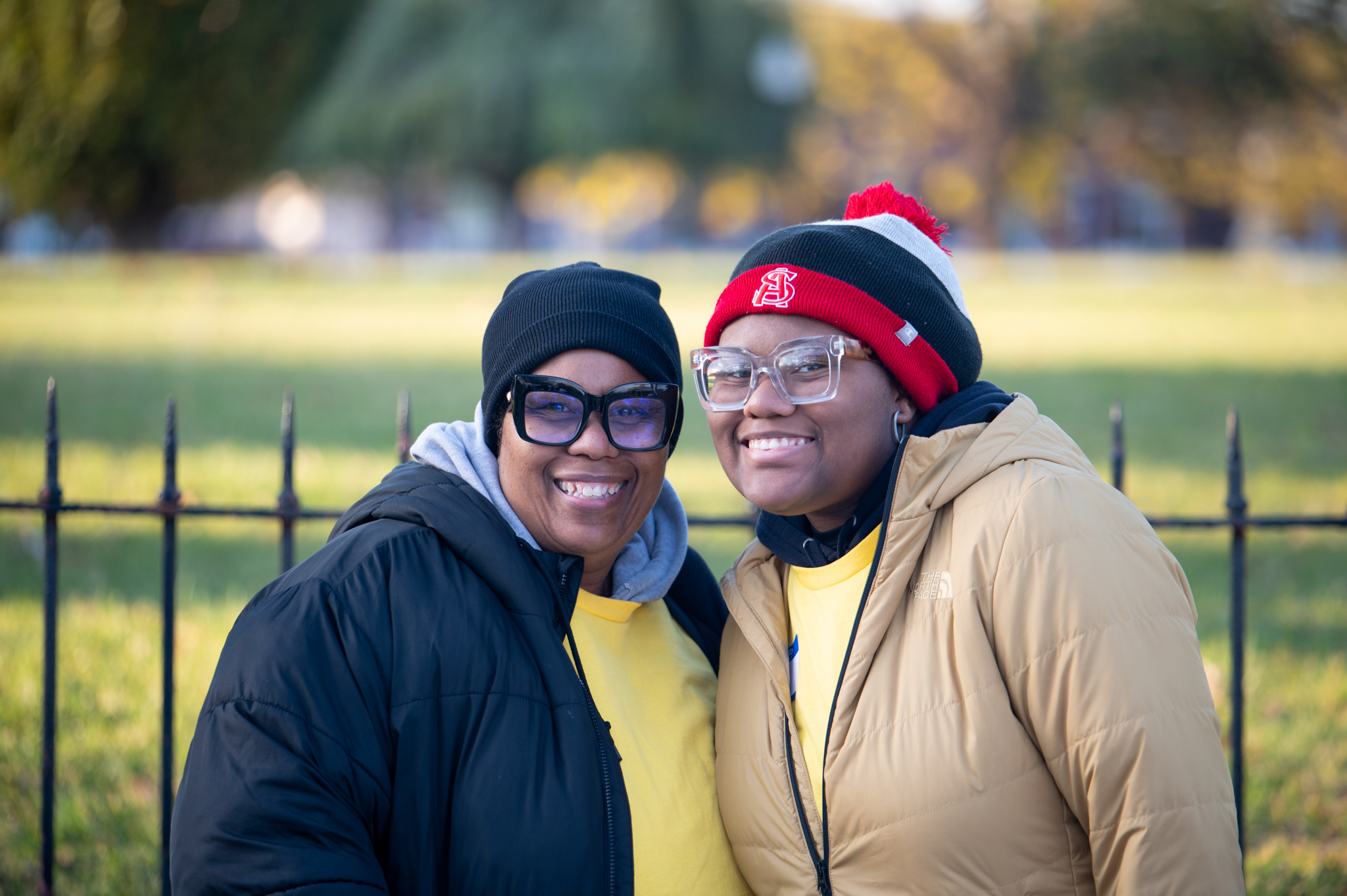 A dark-skinned woman in a black coat and a dark-skinned woman in a tan coat smile together.