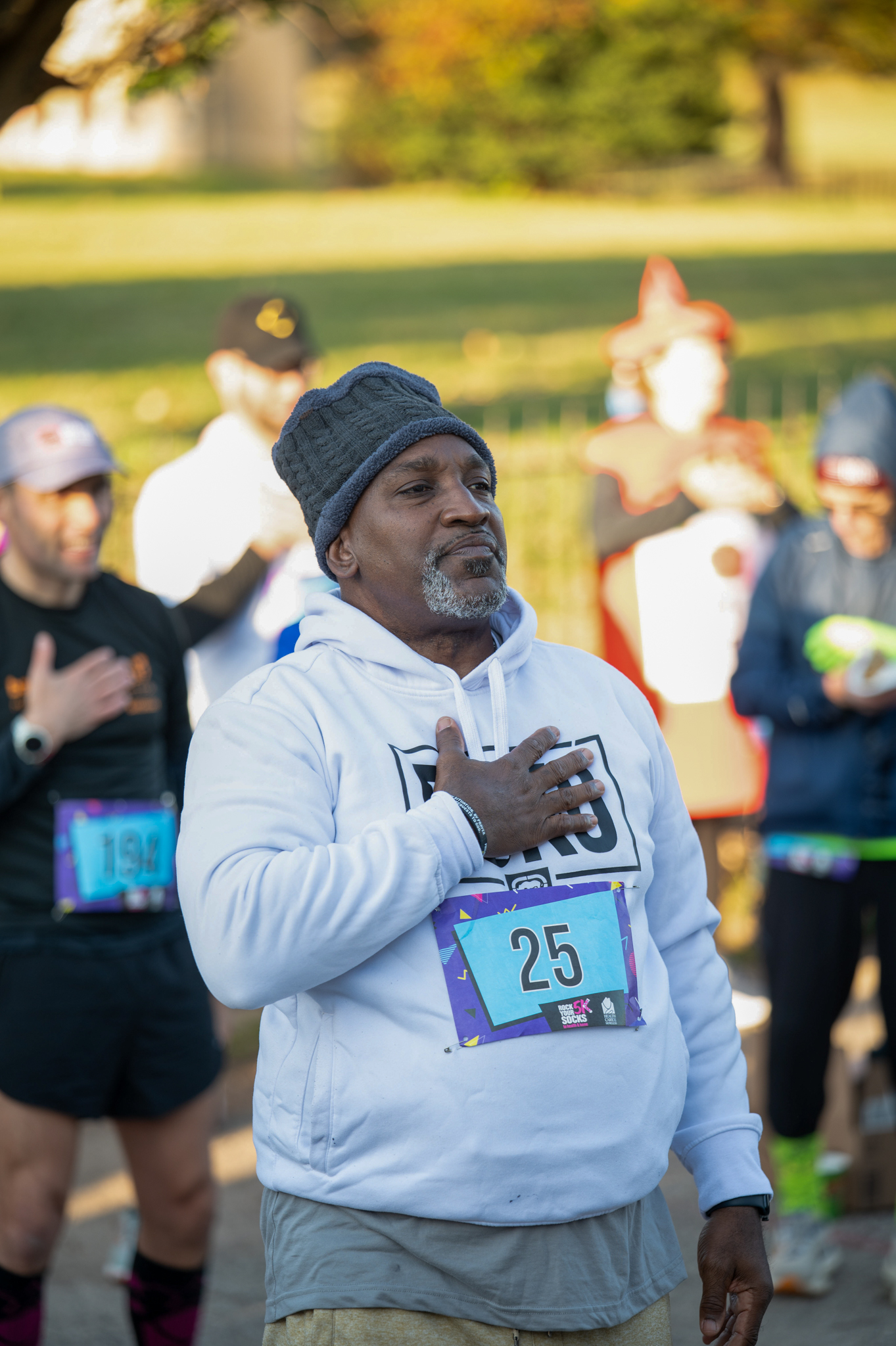 A dark-skinned man wearing a light grey hoodie rests his hand on his chest.