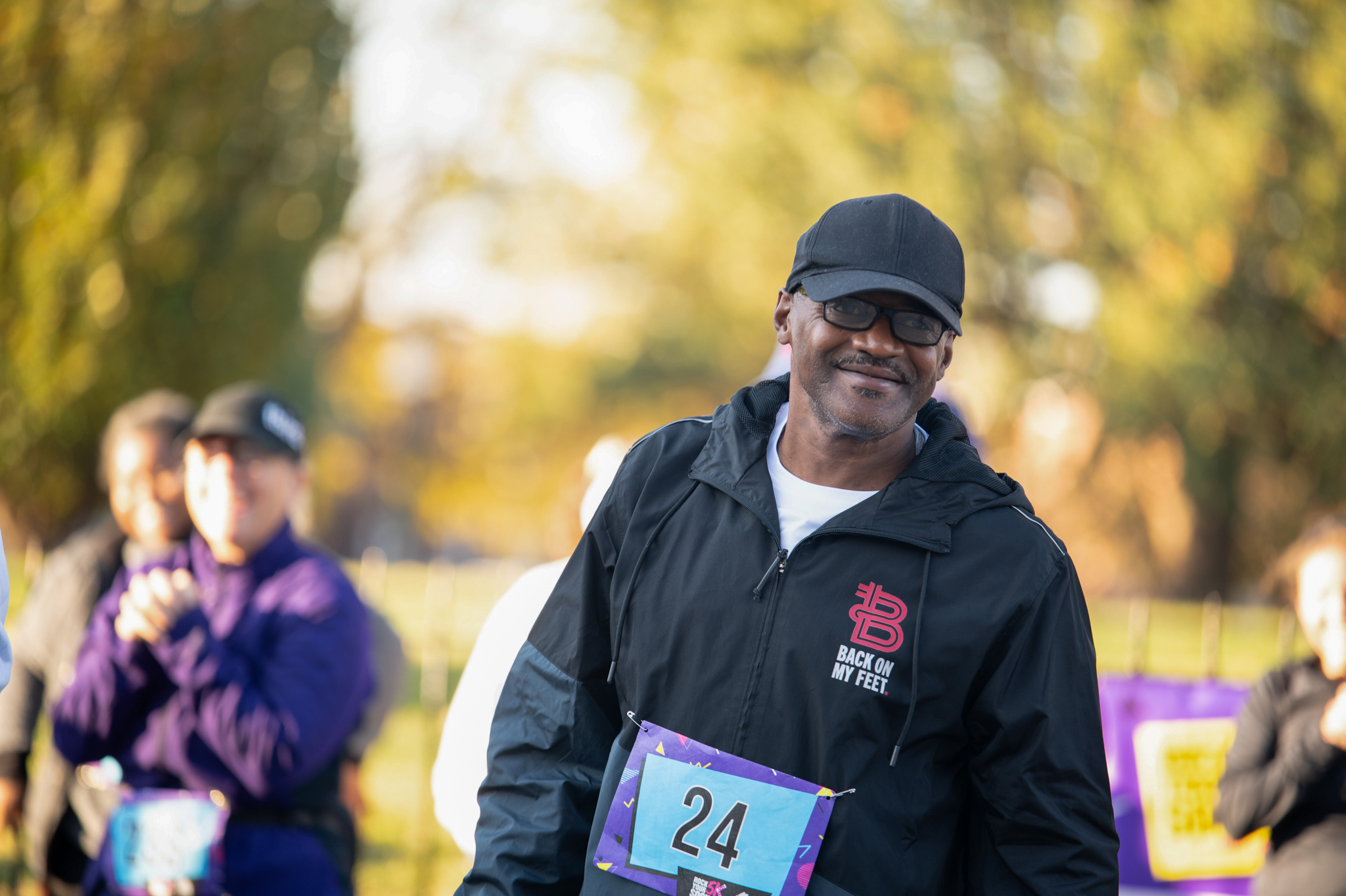 A dakr-skinned man wearing a black windbreaker and black hat smiles.