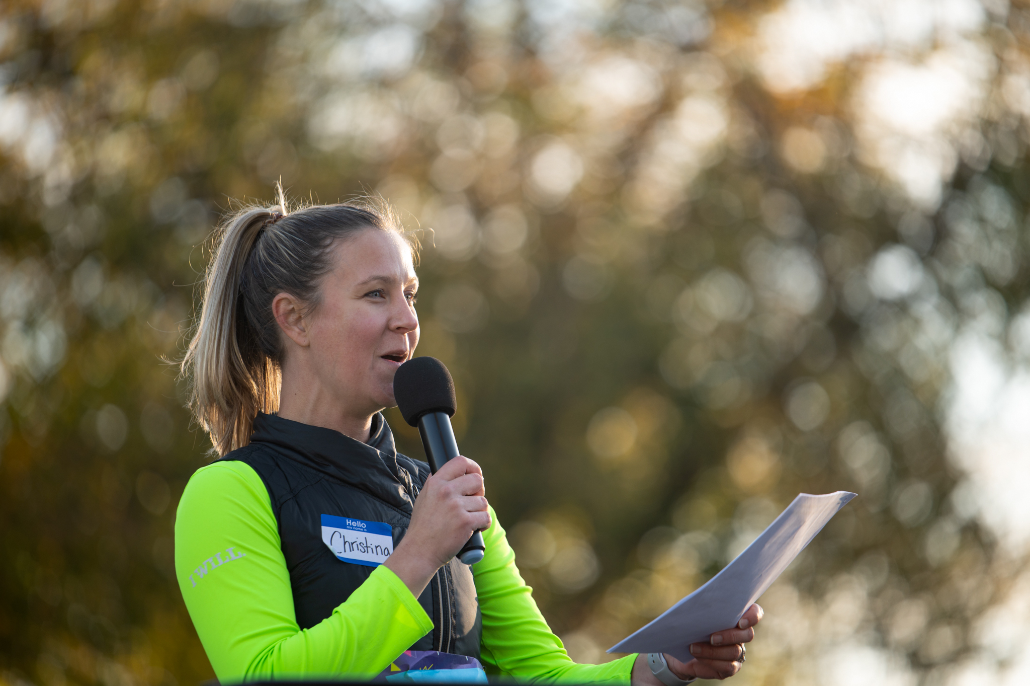 A light-skinned woman holds a microphone and a piece of paper