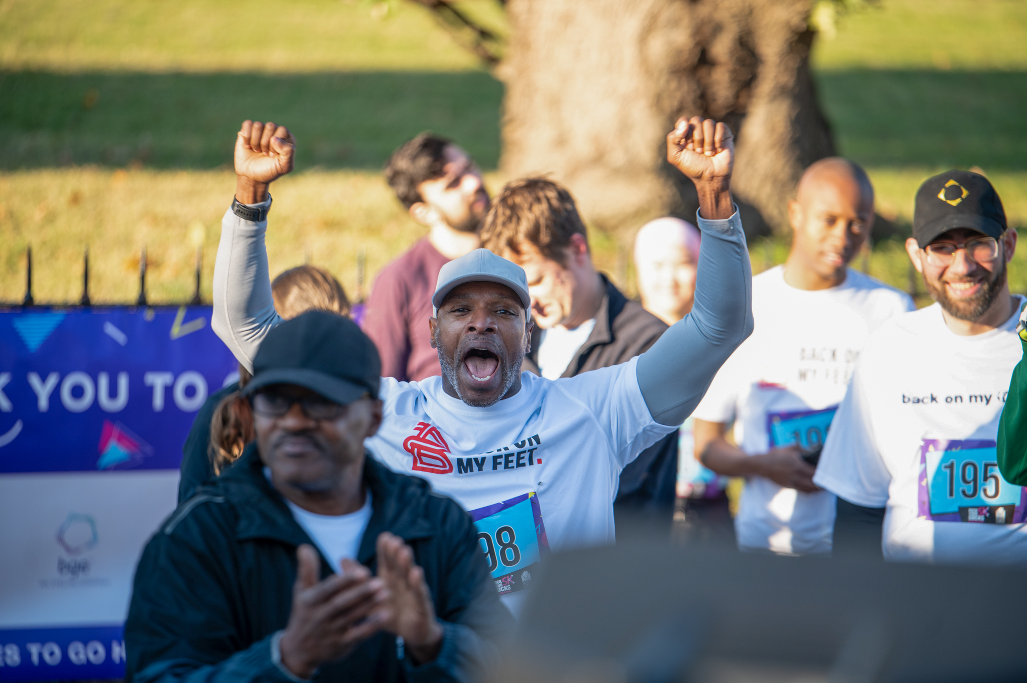 A dark-skinned man wearing a grey hat and a white t-shirt over a grey long-sleeve shirt raises his fists.
