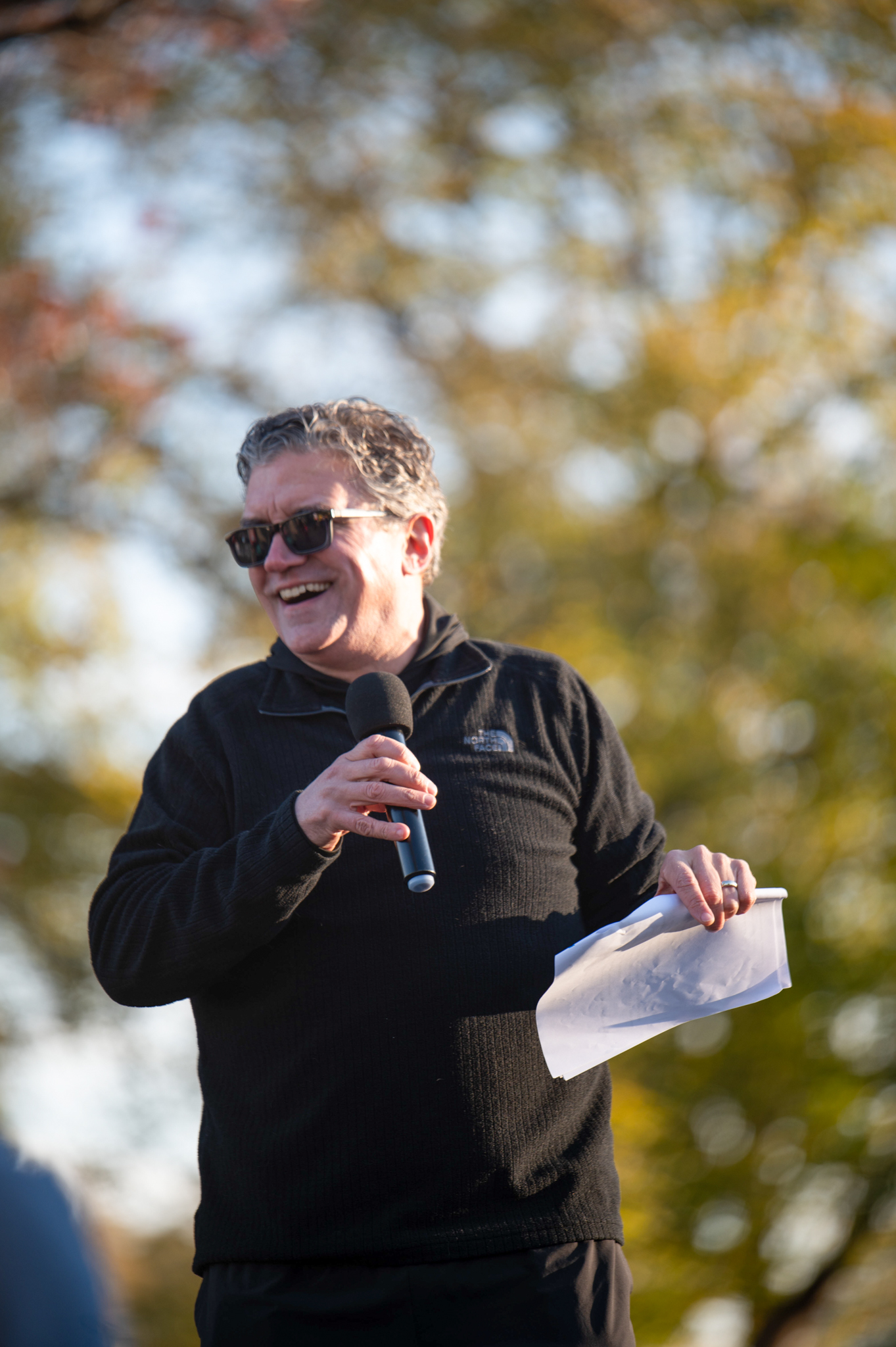 A light-skinned man in a black athletic top hold a microphone and a piece of paper.