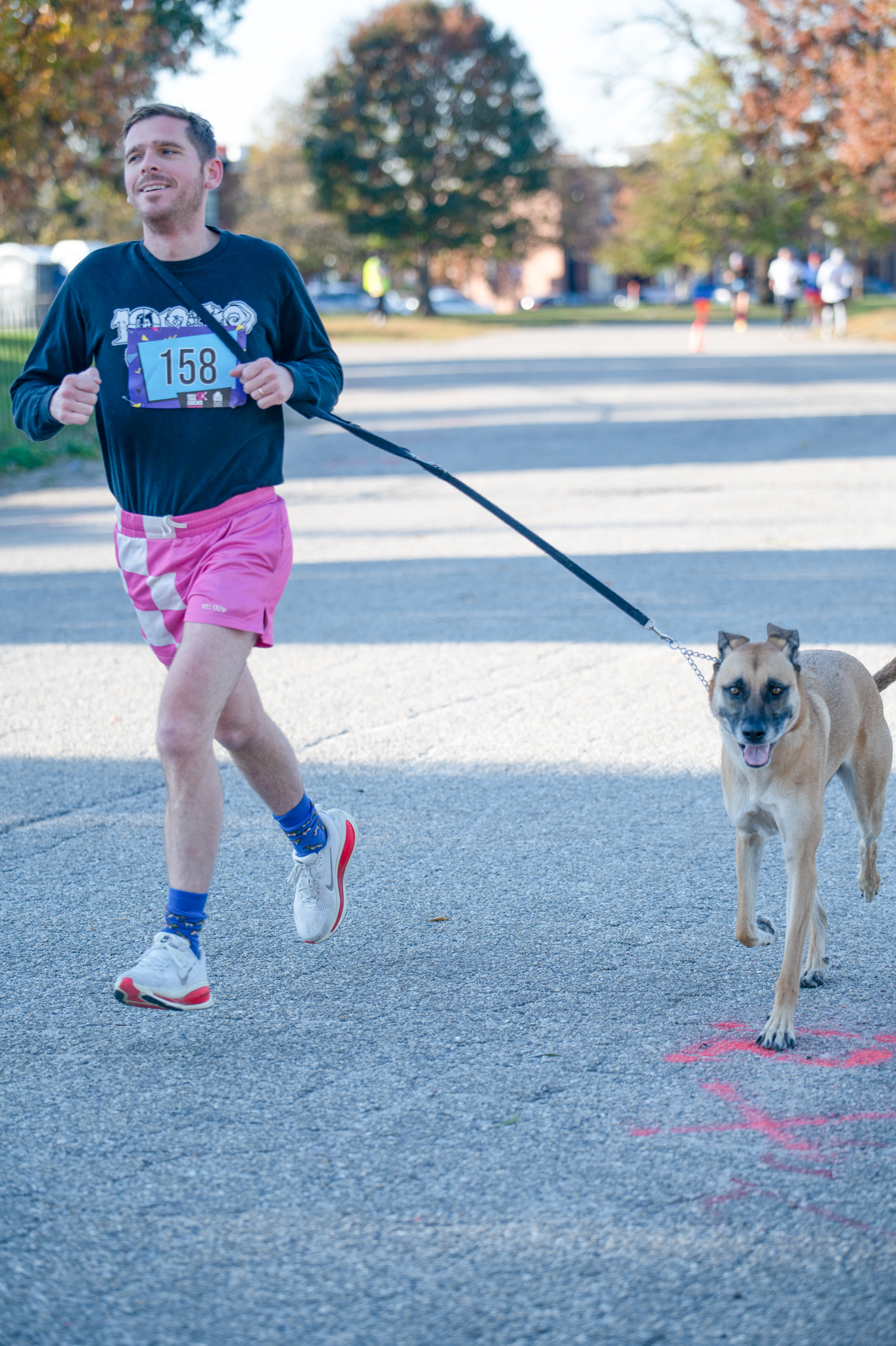A light-skinned man runs in a black shirt and pink shirts with a tan dog