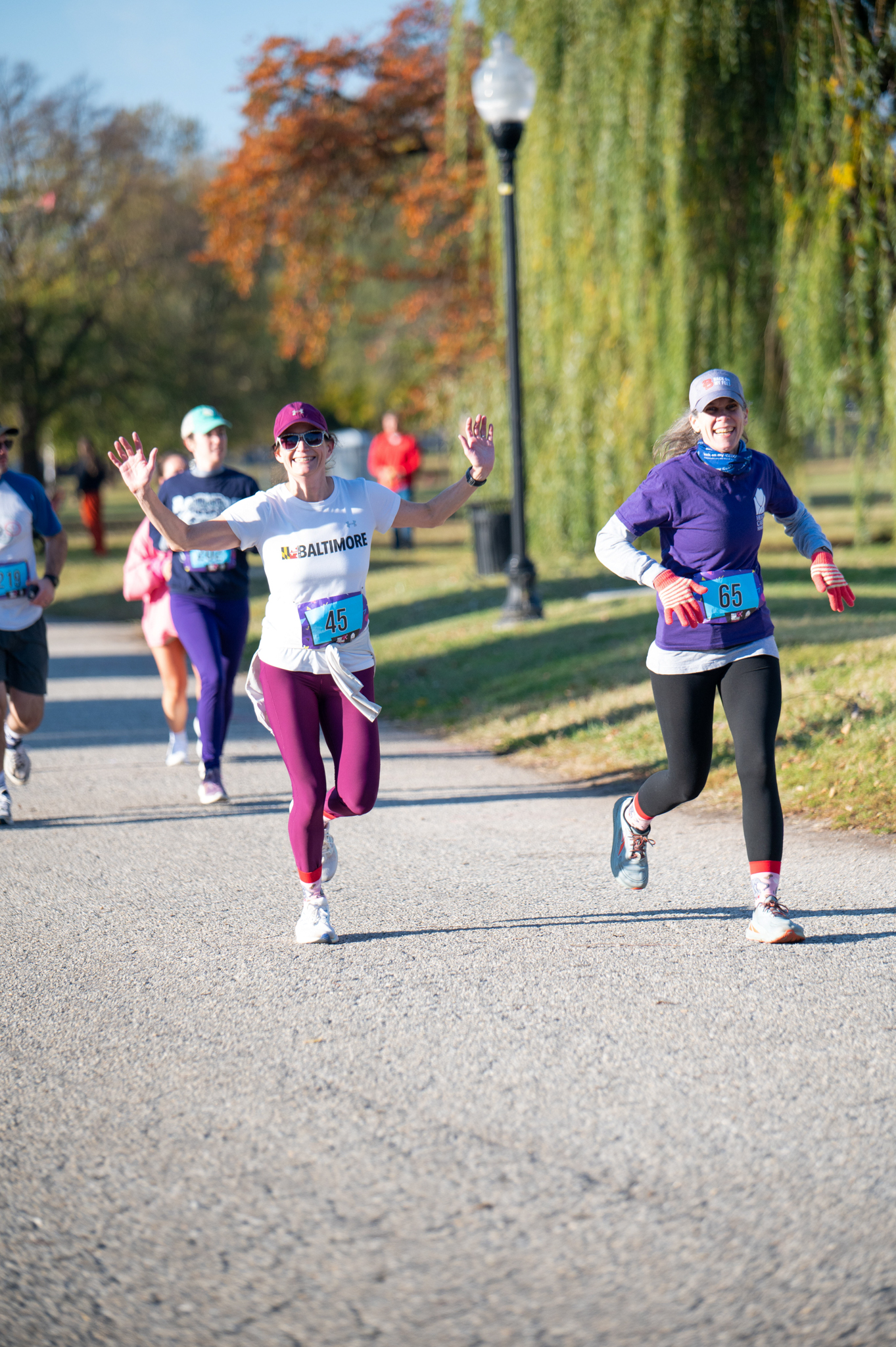 A group of 5K runners. A light-skinned woman in a white top and purple leggings extends her arms.