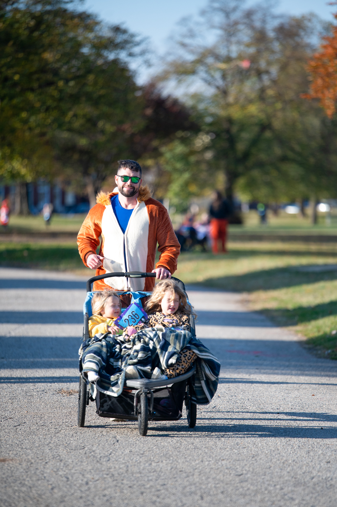 A light-skinned man in a lion costume pushes a double stroller with two girls