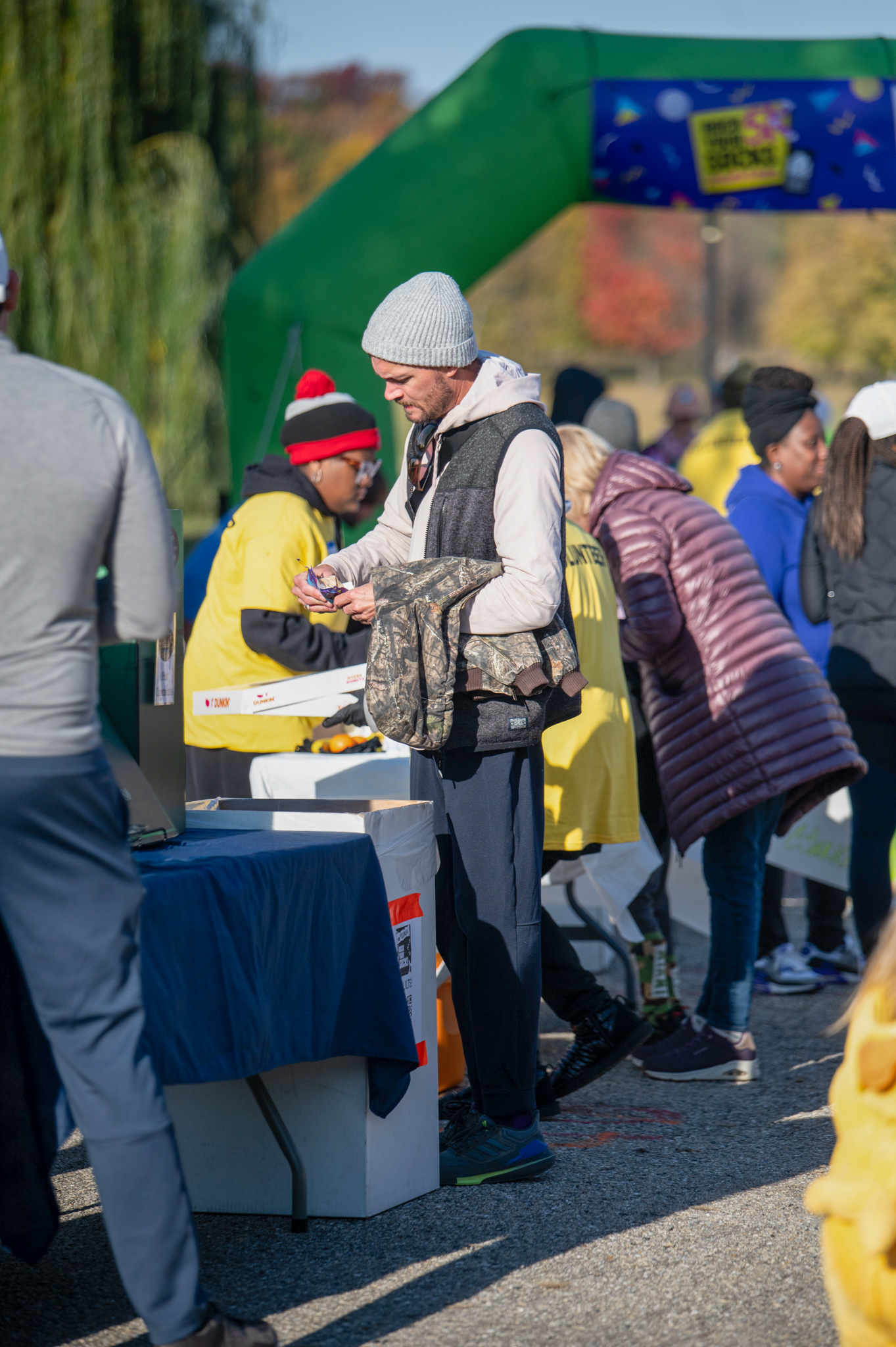 A light-skinned man in a grey hat and black vest stands at a vendor table