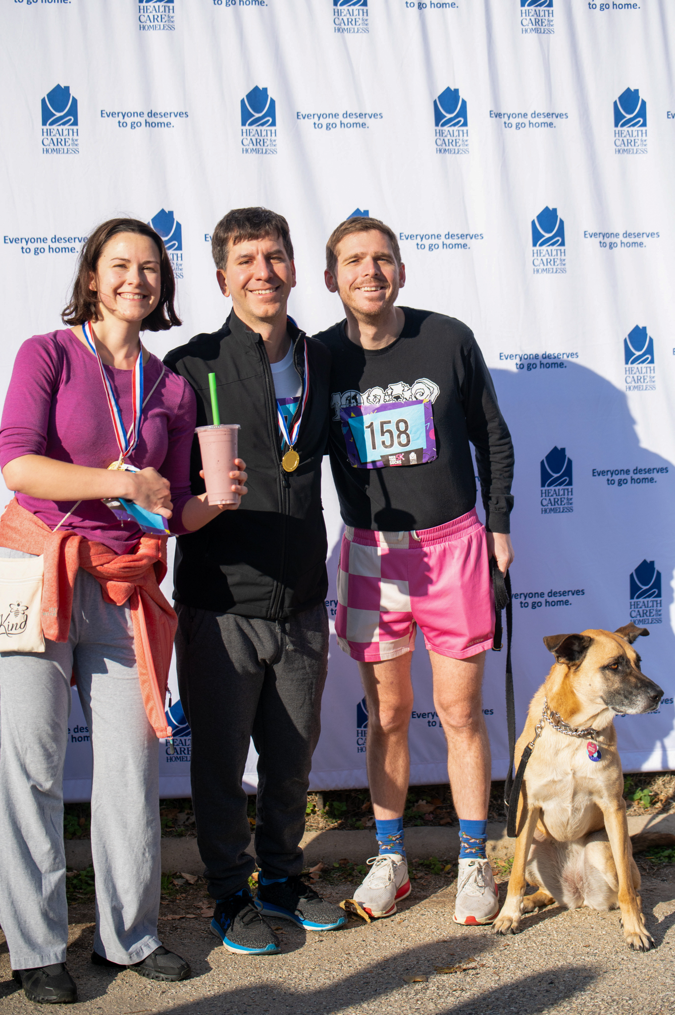 Three light-skinned people smile next to a tan dog in front of the step-and-repeat