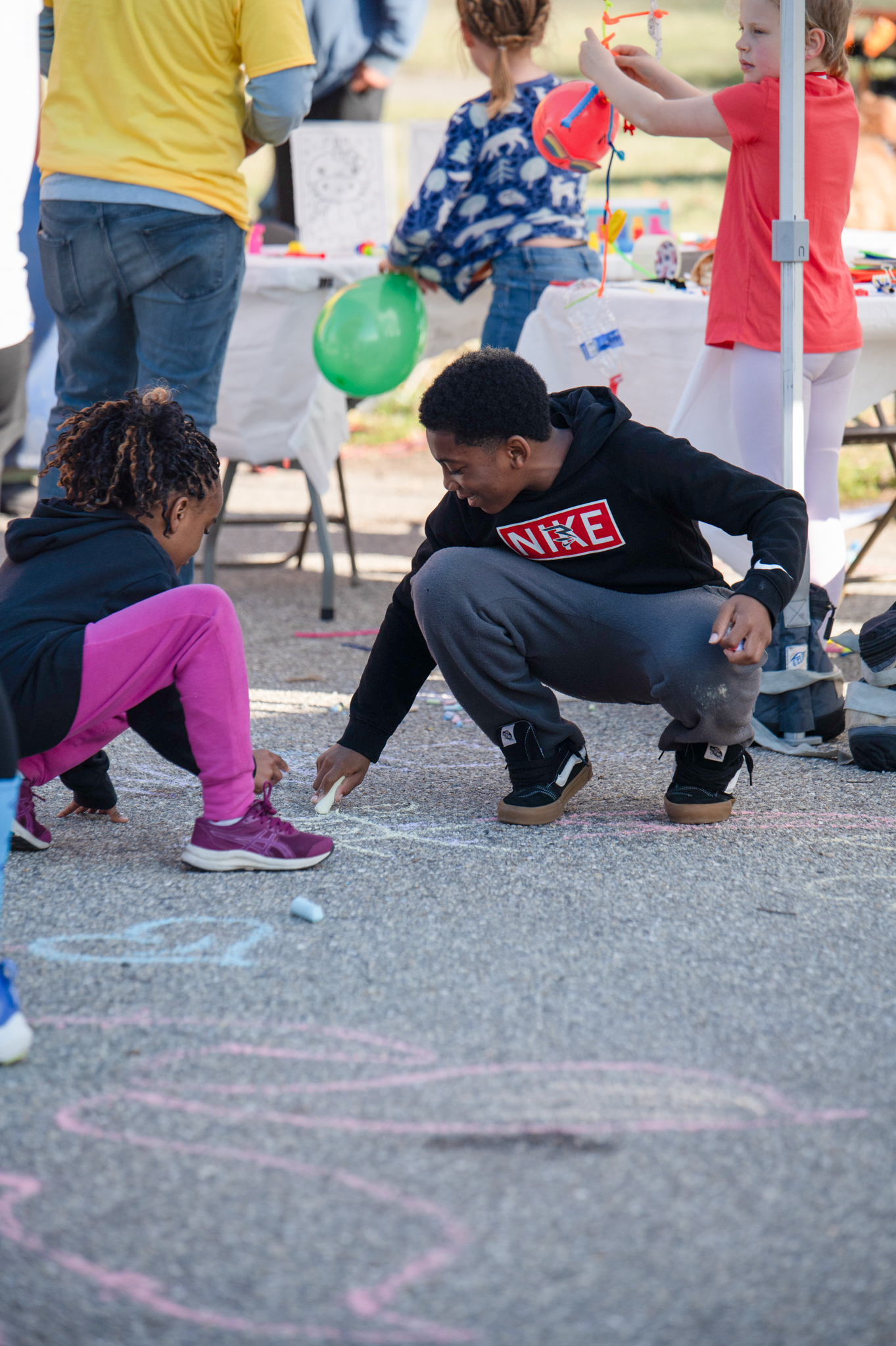 A dark-skinned girl in pink pants draws with chalk with a dark-skinned boy wearing a black hoodie