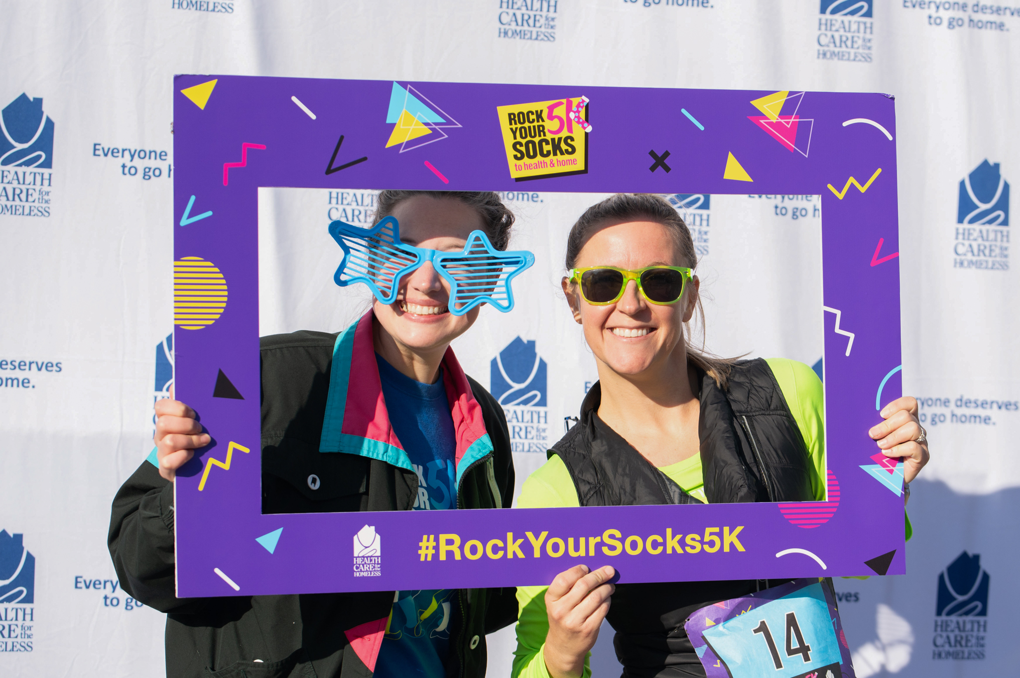 Two light-skinned women smile while holding a purple photo frame in front of the Health Care for the Homeless step-and-repeat