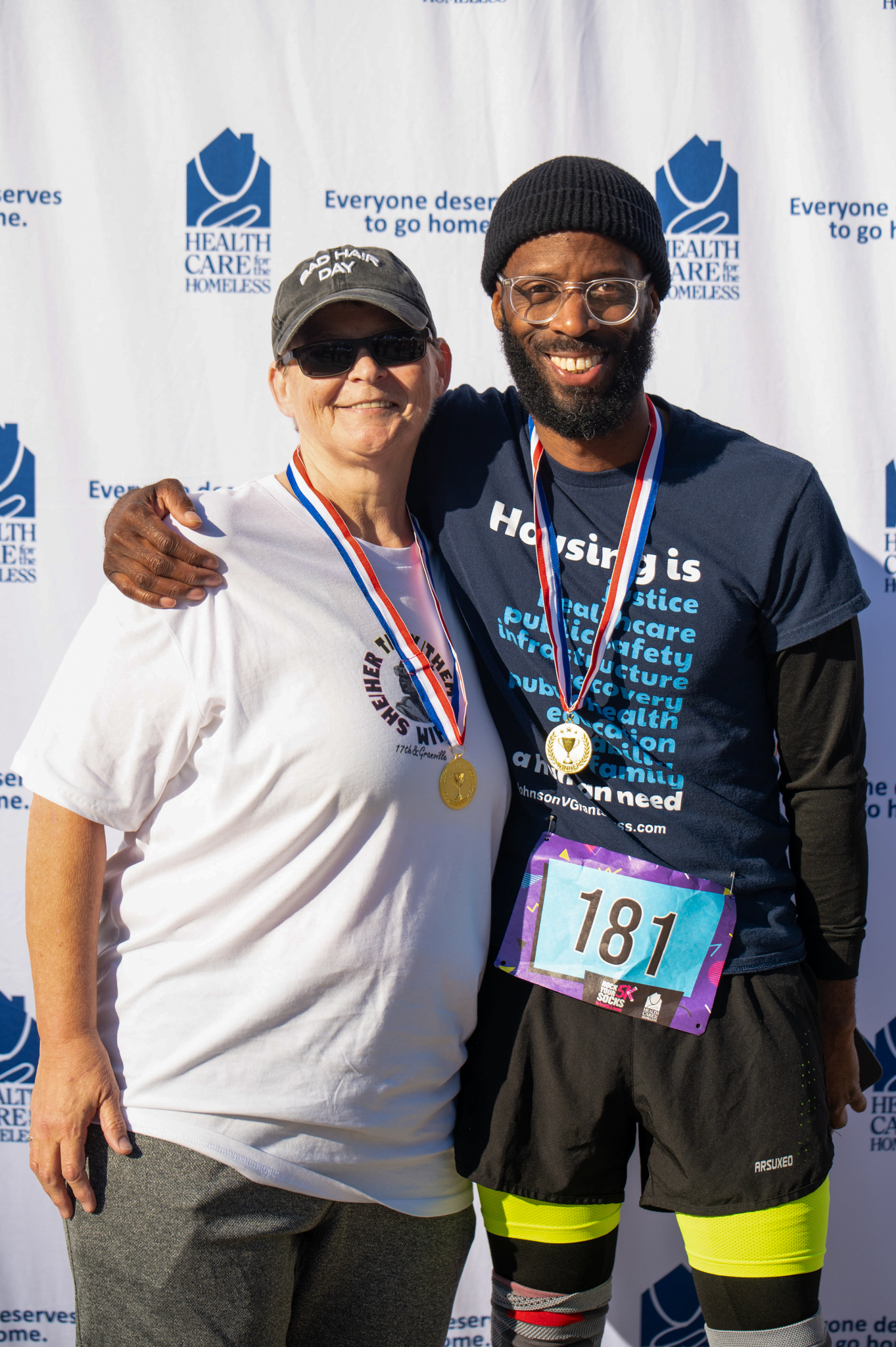 A light-skinned person wearing a hat and sunglasses poses with a dark-skinned man wearing a hat and medal in front of the Health Care for the Homeless step-and-repeat