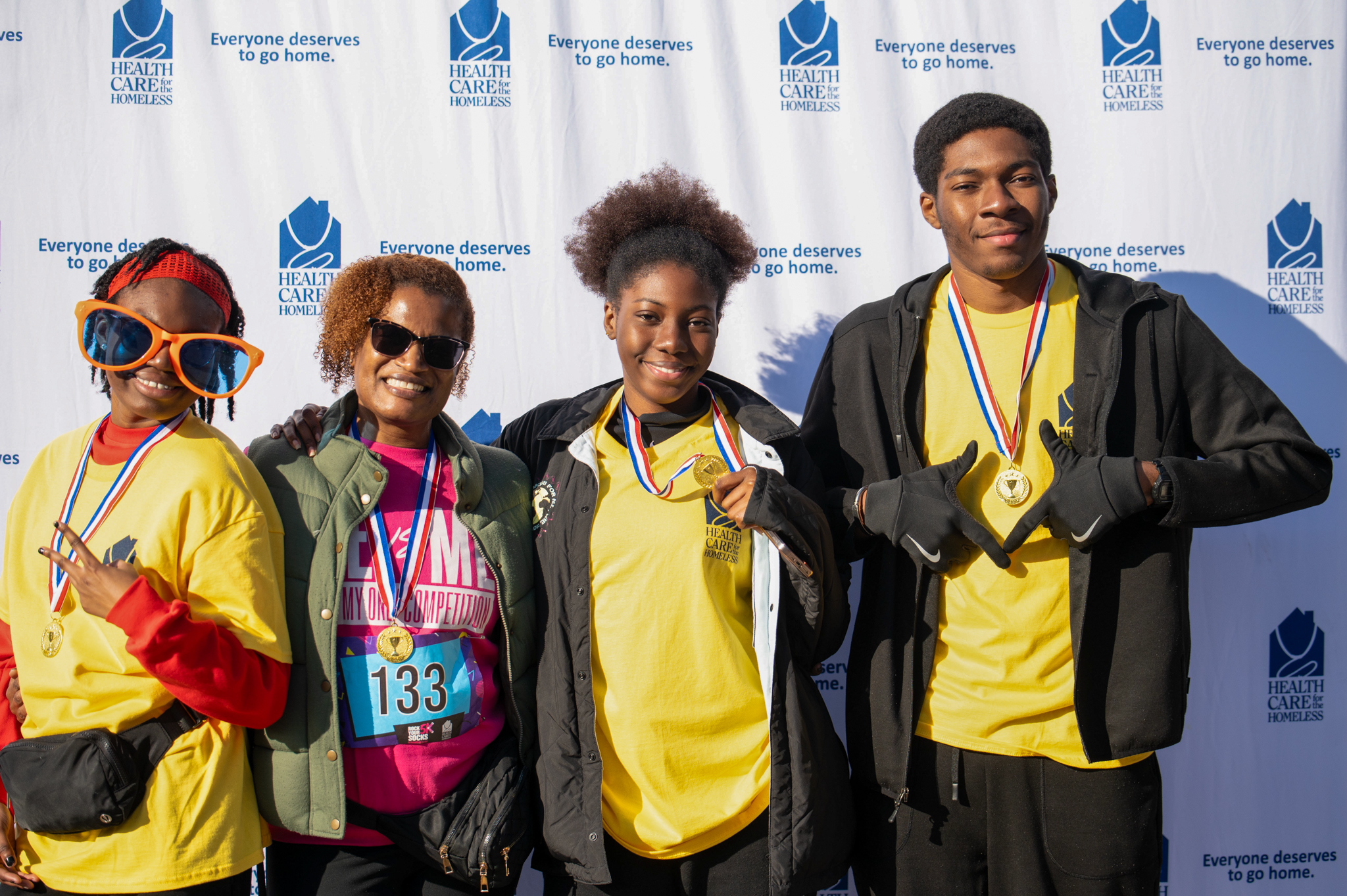 A group of four hold 80s props and pose in front of the Health Care for the Homeless step-and-repeat
