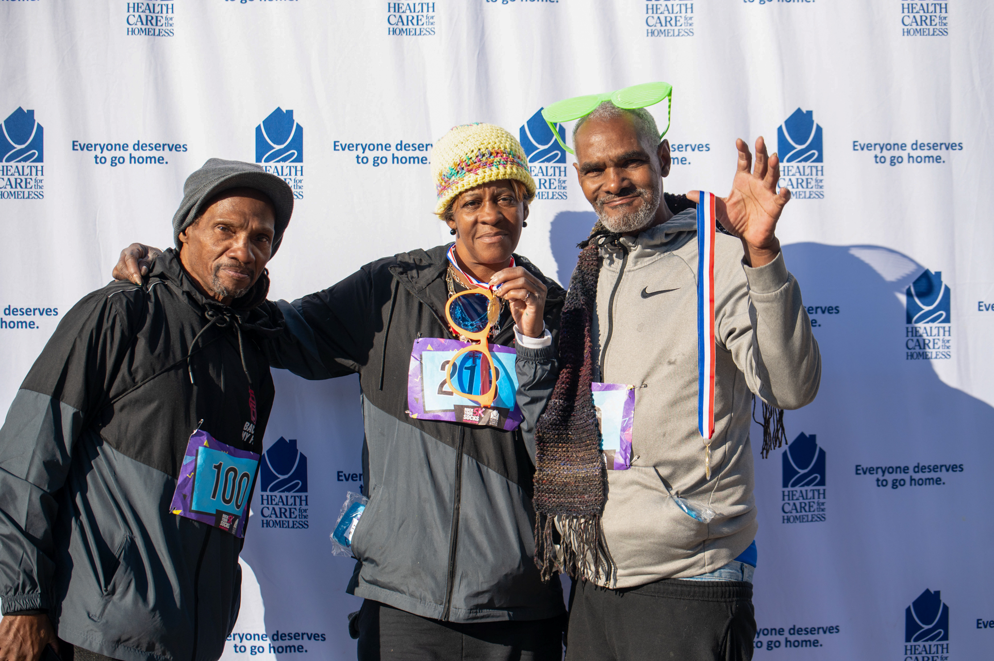 A group holding medals and 80s props stand in front of the Health Care for the Homeless step-and-repeat