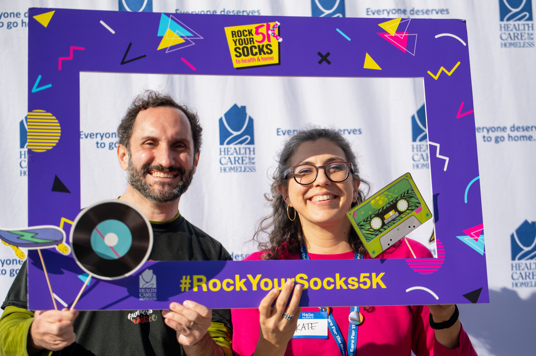 A light-skinned man in a black top and a light-skinned woman in punk top hold a purple photo frame