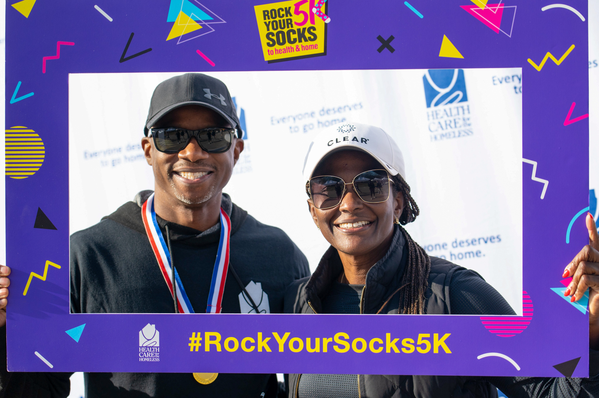 A dark-skinned man in a black hat and a dark-skinned woman in a white hat hold a purple photo frame