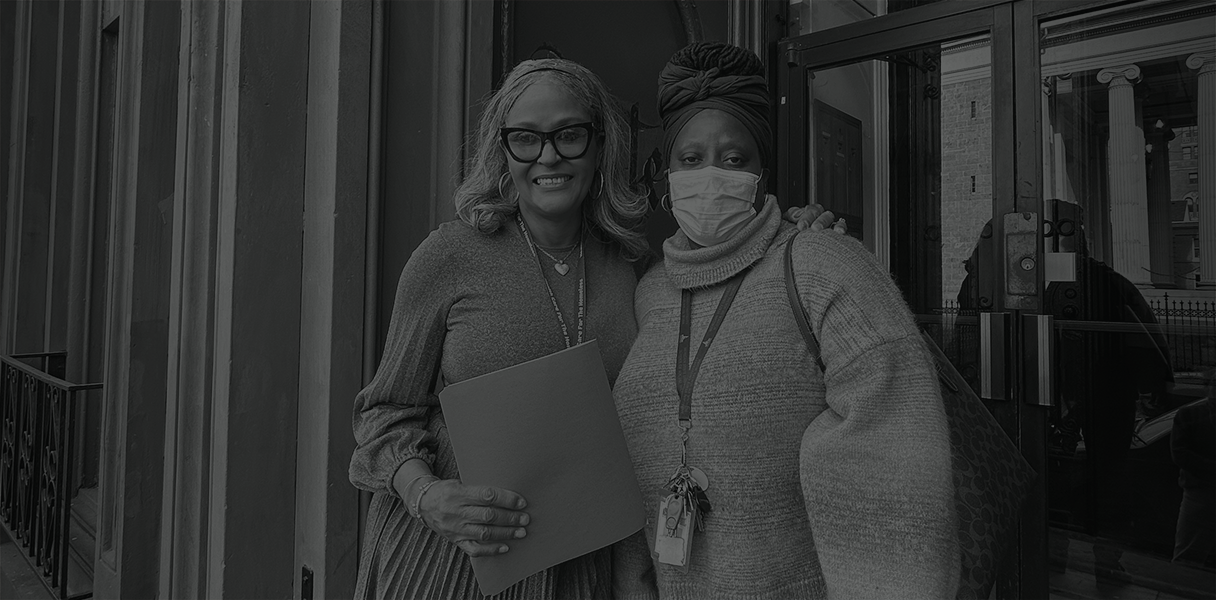 A woman with glasses and a woman wearing a mask stand in front of Mulberry Supportive Housing.