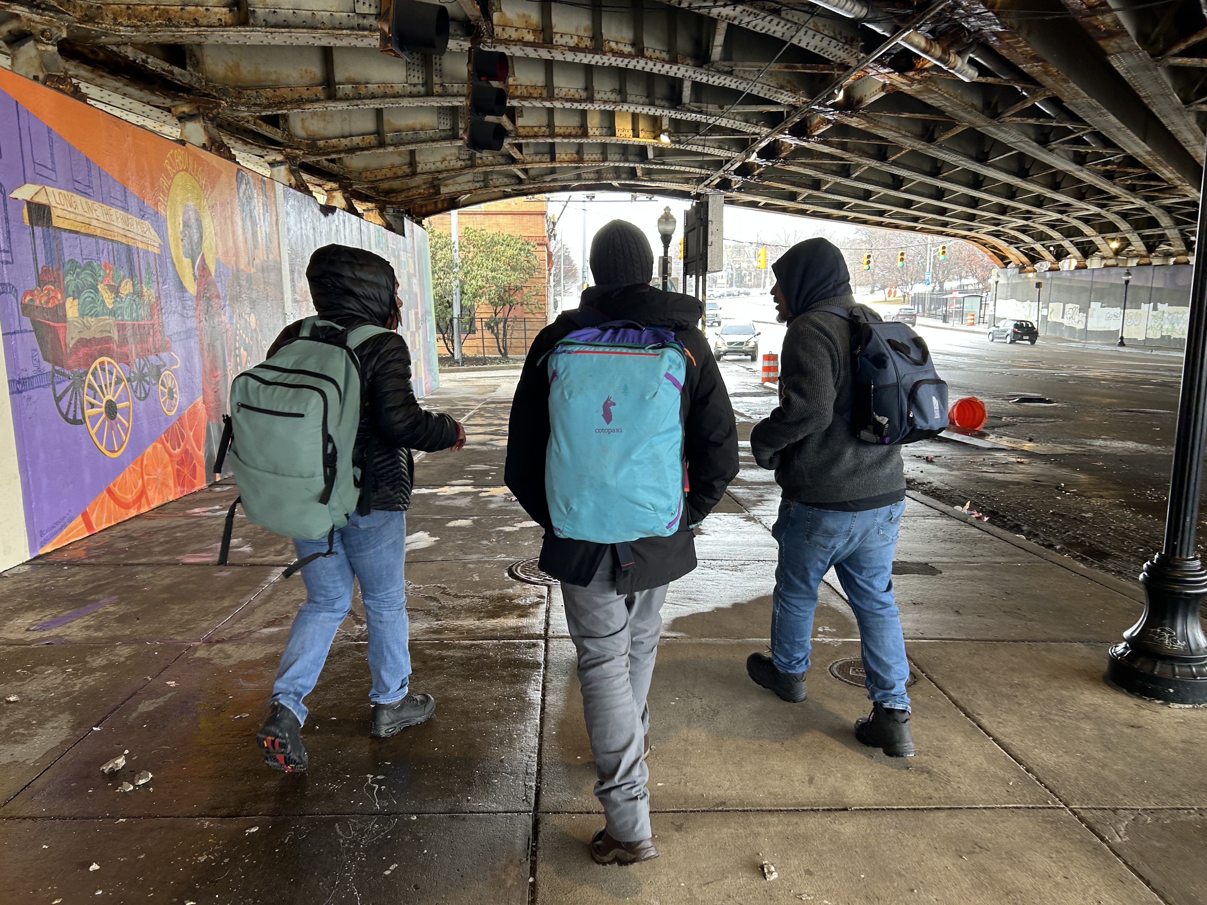 Three people wear large backpacks, backs toward camera, under an overpass.
