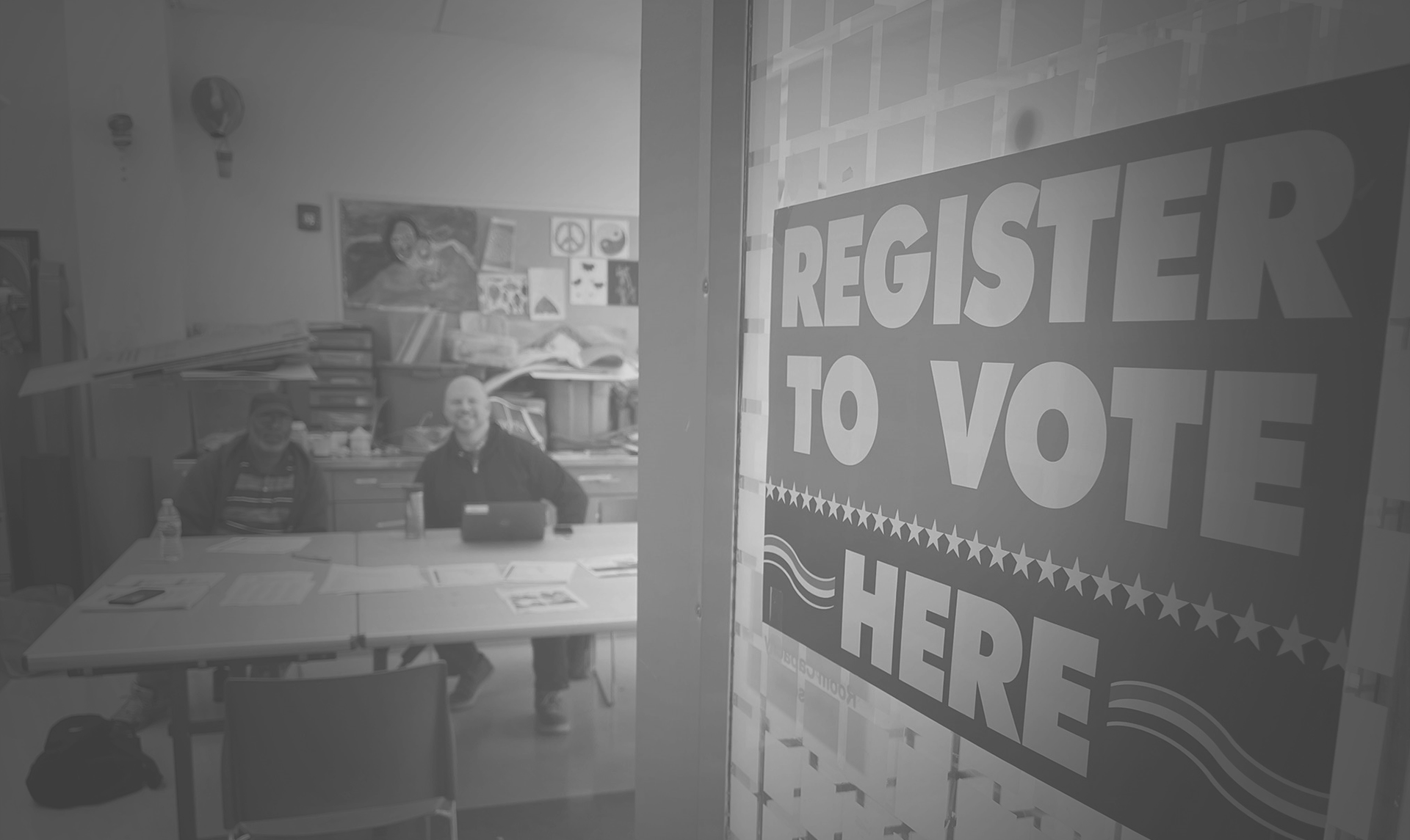 Black and white image of two people sitting at a table. In the forefront is a sign that reads, "Register to vote here."