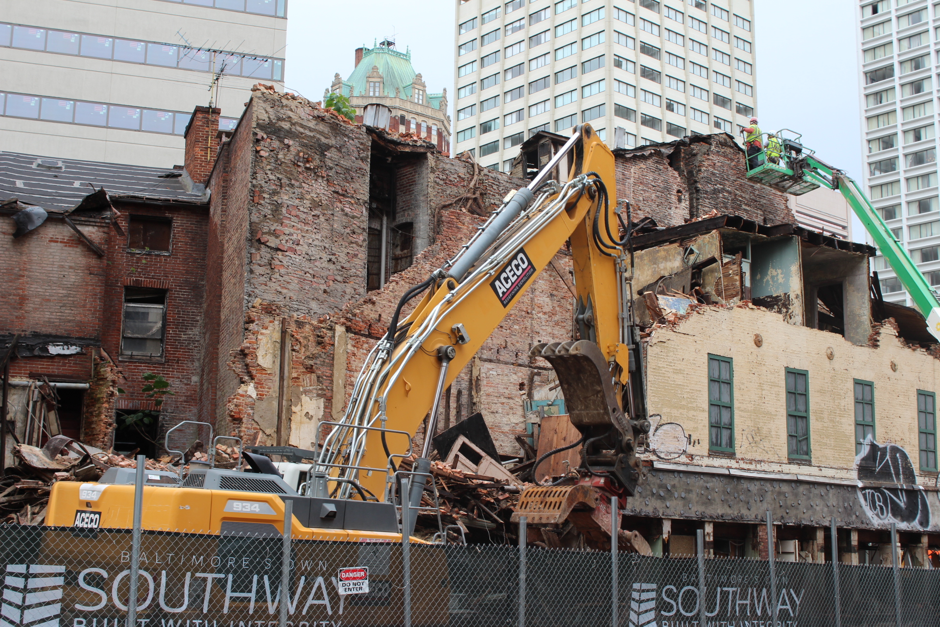 A yellow excavator moves building debris in front of a brick building under construction..