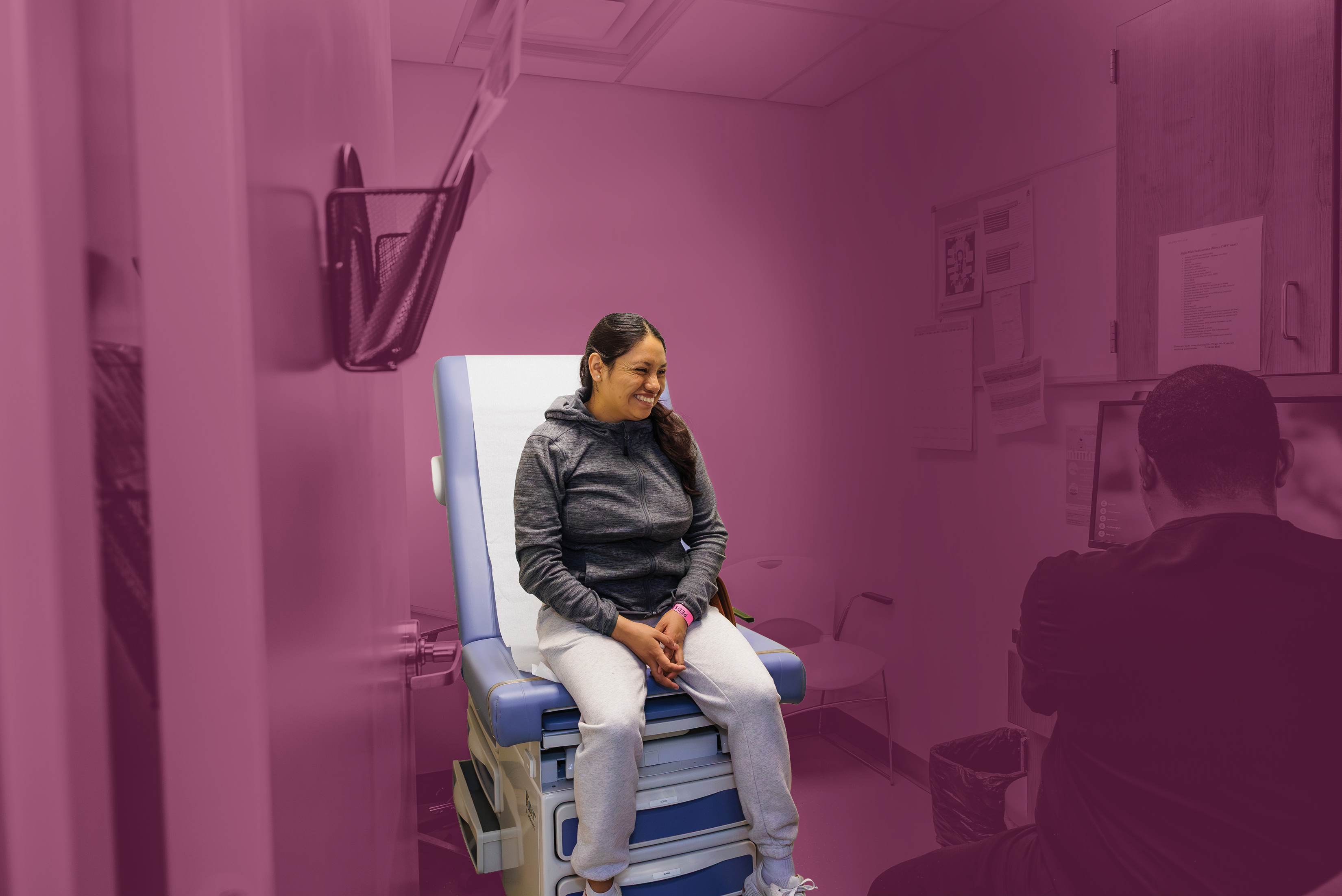 A woman sits on an exam room table. She is in full color, the rest of the room has a magenta filter