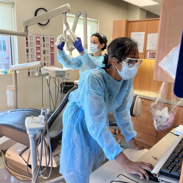 One dental staff member in a blue gown types at a computer while another adjusts equipment.
