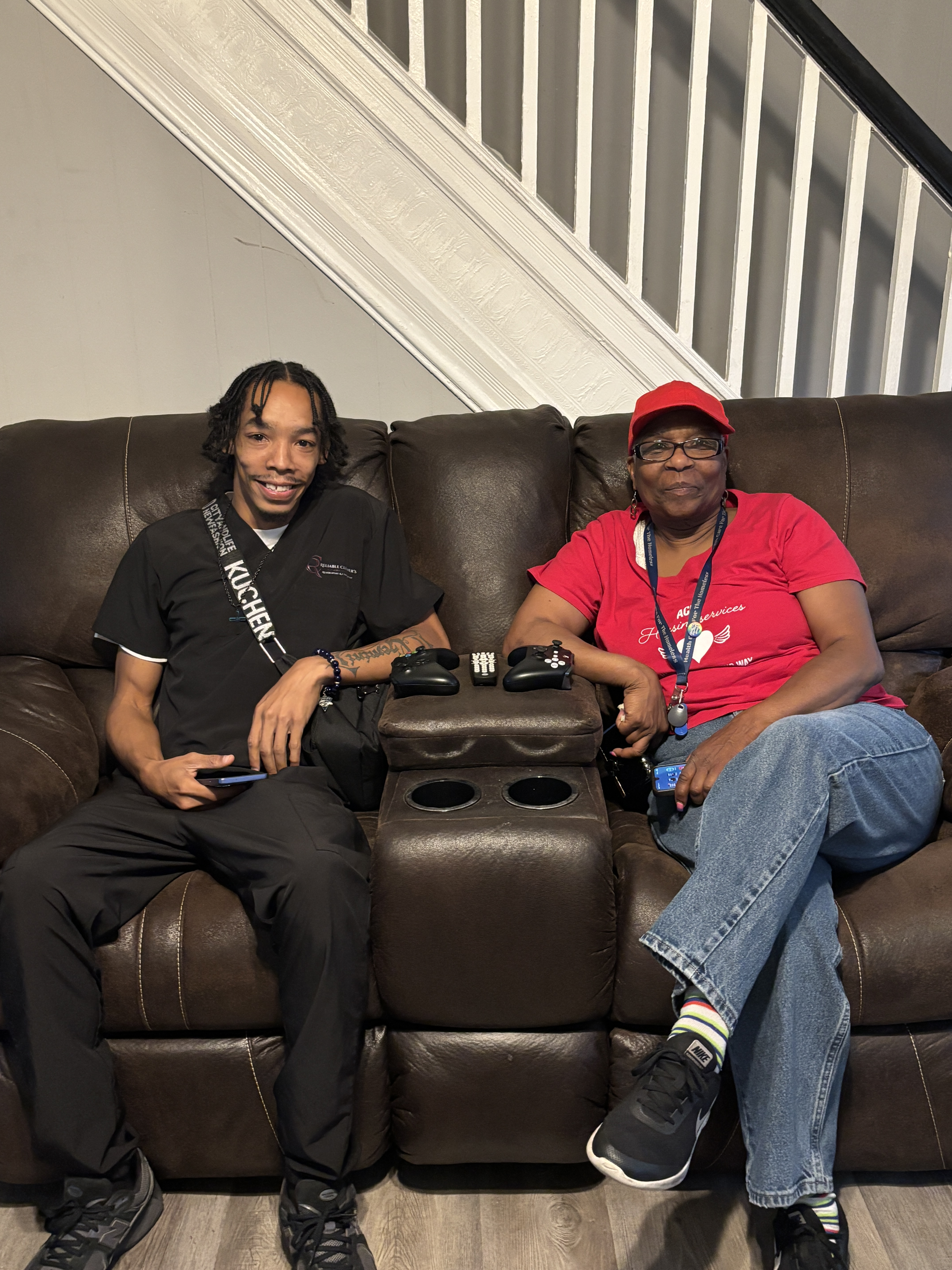 A men in dark clothing sits on a brown couch next to a woman in a red hat and red shirt.