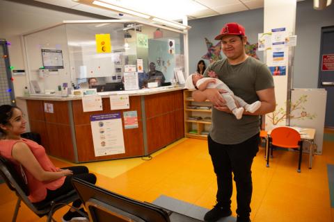 A man wearing a red baseball cap stands in a clinic waiting area cradling his small child