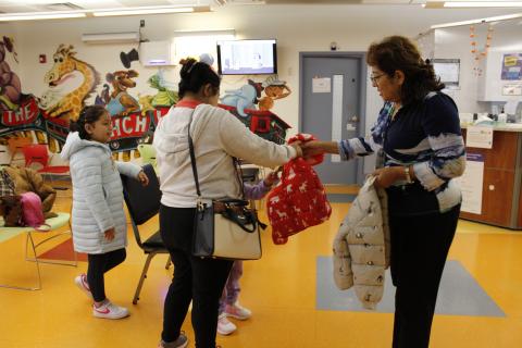 A medium-skinned woman with light brown hair hands a red coat to a dark-haired woman standing with two girls.
