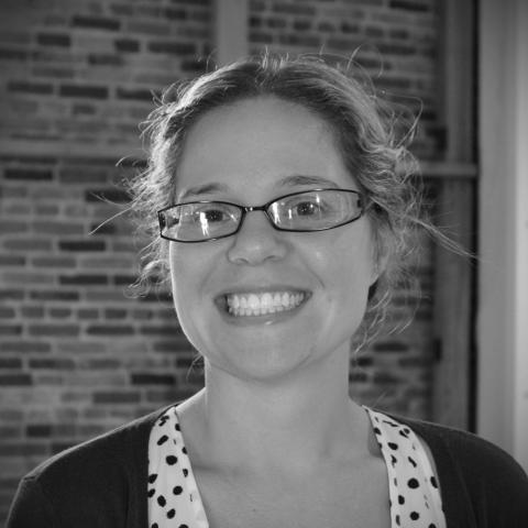 A black and white headshot of a light-skinned woman with glasses and hair pulled back.