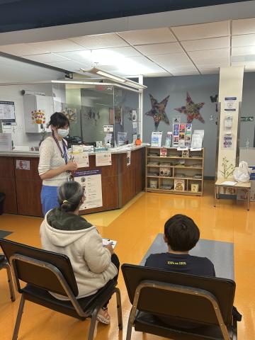 A light-skinned woman speaks to a mother and son sitting in chairs in the waiting room of the Pediatric clinic
