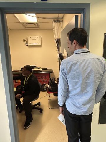 A dark-skinned man sits on a stool and a light-skinned man faces him from a doorway