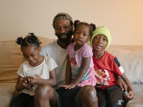 A dark-skinned man holds his two daughters and son in his lap on a couch